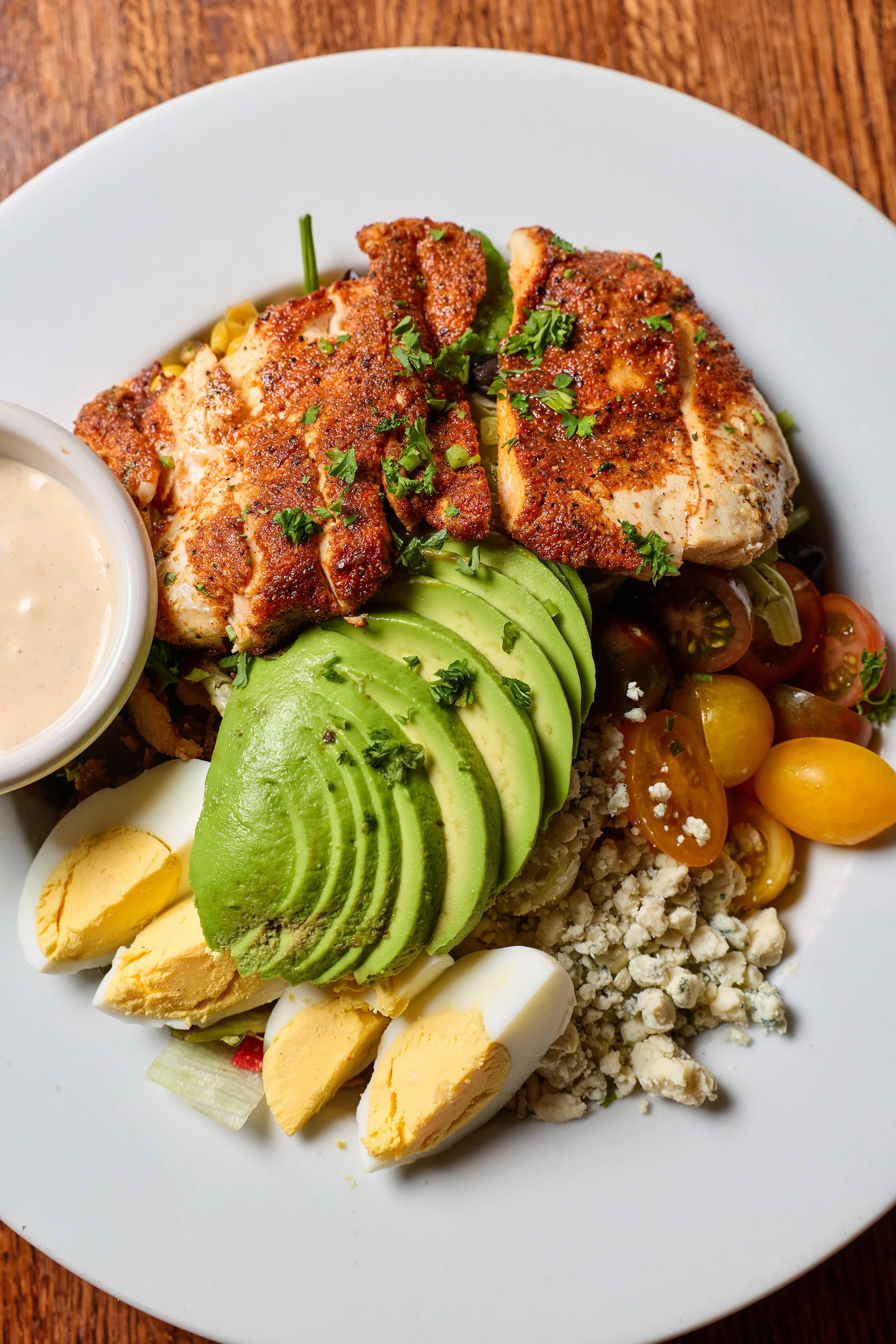 Close-up of a breakfast plate with sliced avocado, sliced hard-boiled eggs, crumbled blue cheese, cherry tomatoes, and grilled chicken breast garnished with chopped herbs, with a small cup of sauce on the side.