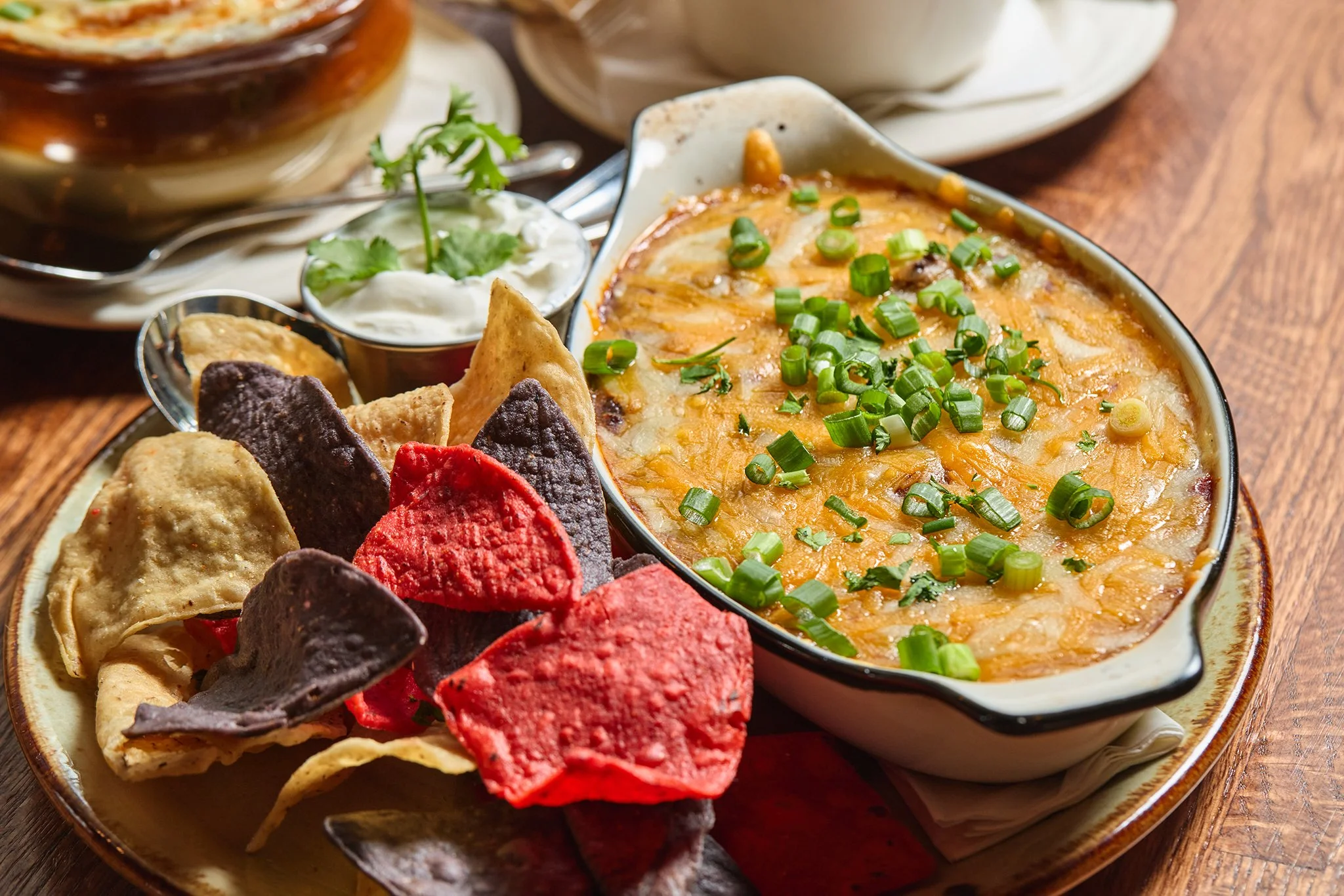 A dish of baked cheese with chopped green onions, garnished with cilantro, served with a side of colorful tortilla chips on a wooden table.