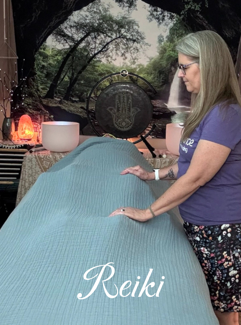 A woman with long gray hair and glasses is giving a Reiki healing session, placing her hands on a person lying on a table covered with a textured blue cloth. The setting includes crystals, a salt lamp, a large bowl, and a decorative backdrop with a nature scene.