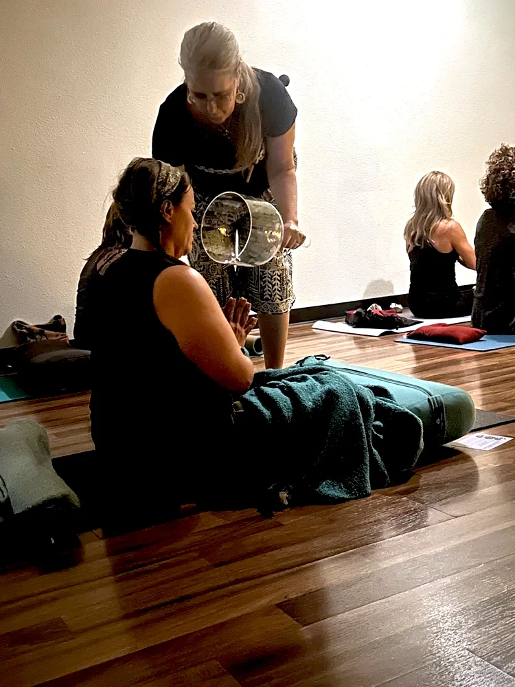 A woman in a yoga class receiving instruction from the teacher. The woman is sitting on a yoga mat with a towel draped over her legs. Other women are practicing yoga in the background.