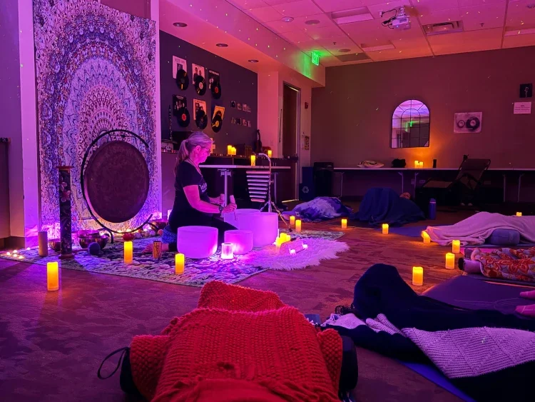 A woman playing a singing bowl in a dimly lit room decorated with candles and soft lighting, with people lying on mats covered in blankets, engaging in a relaxation or meditation session.
