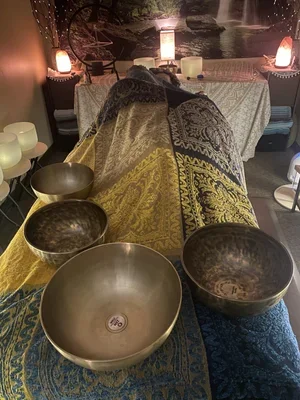 A row of singing bowls placed on colorful fabric, with a cozy room in the background featuring dim lighting, lamps, and furniture.
