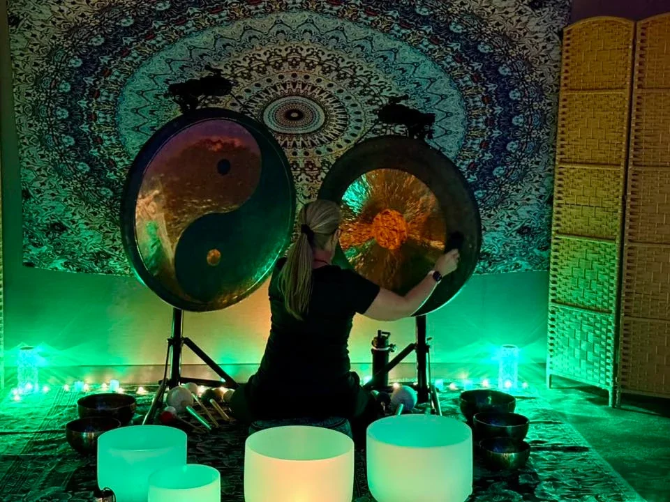 A woman seated on a cushion playing large gong instruments in a dimly lit room with green lighting, surrounded by bowls and decorative items, and a colorful tapestry with a mandala pattern on the wall.