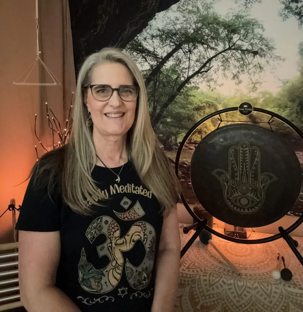 A smiling woman with long blonde hair and glasses standing indoors in front of a nature-themed mural. She wears a black t-shirt with spiritual symbols and a necklace. Behind her is a large gong with a symbolic hand design, and the room is decorated with fairy lights and a tablet on a stand.