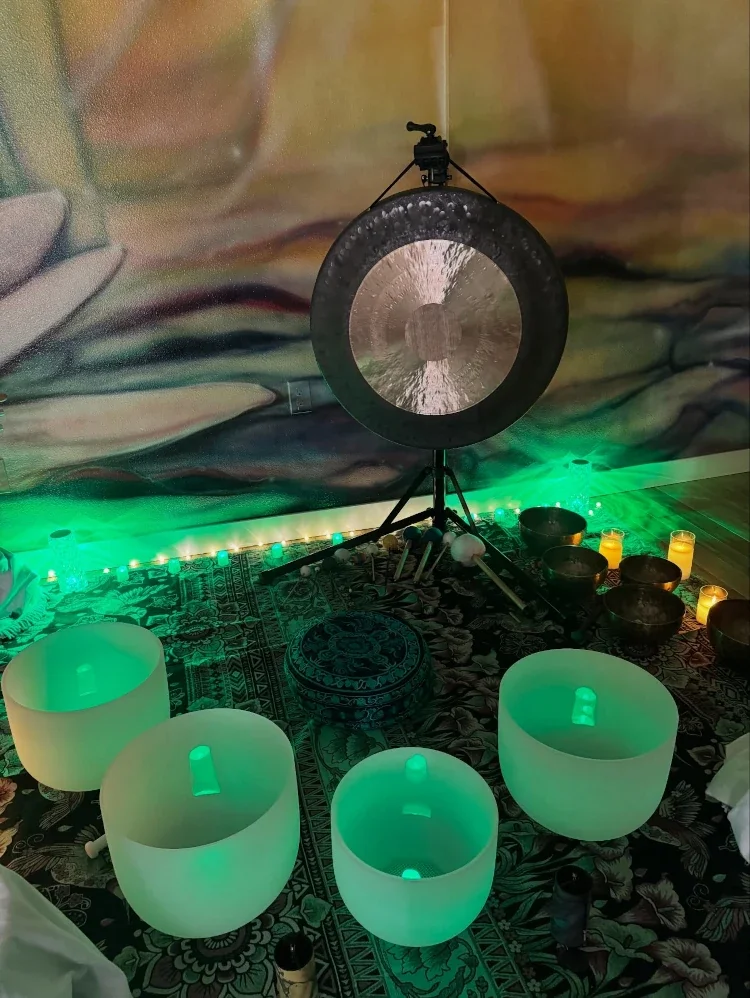 A setup for sound healing or meditation featuring four glowing crystal singing bowls arranged on a decorative cloth, a large gong on a stand in the background, and green candles and LED lights creating a calming atmosphere.