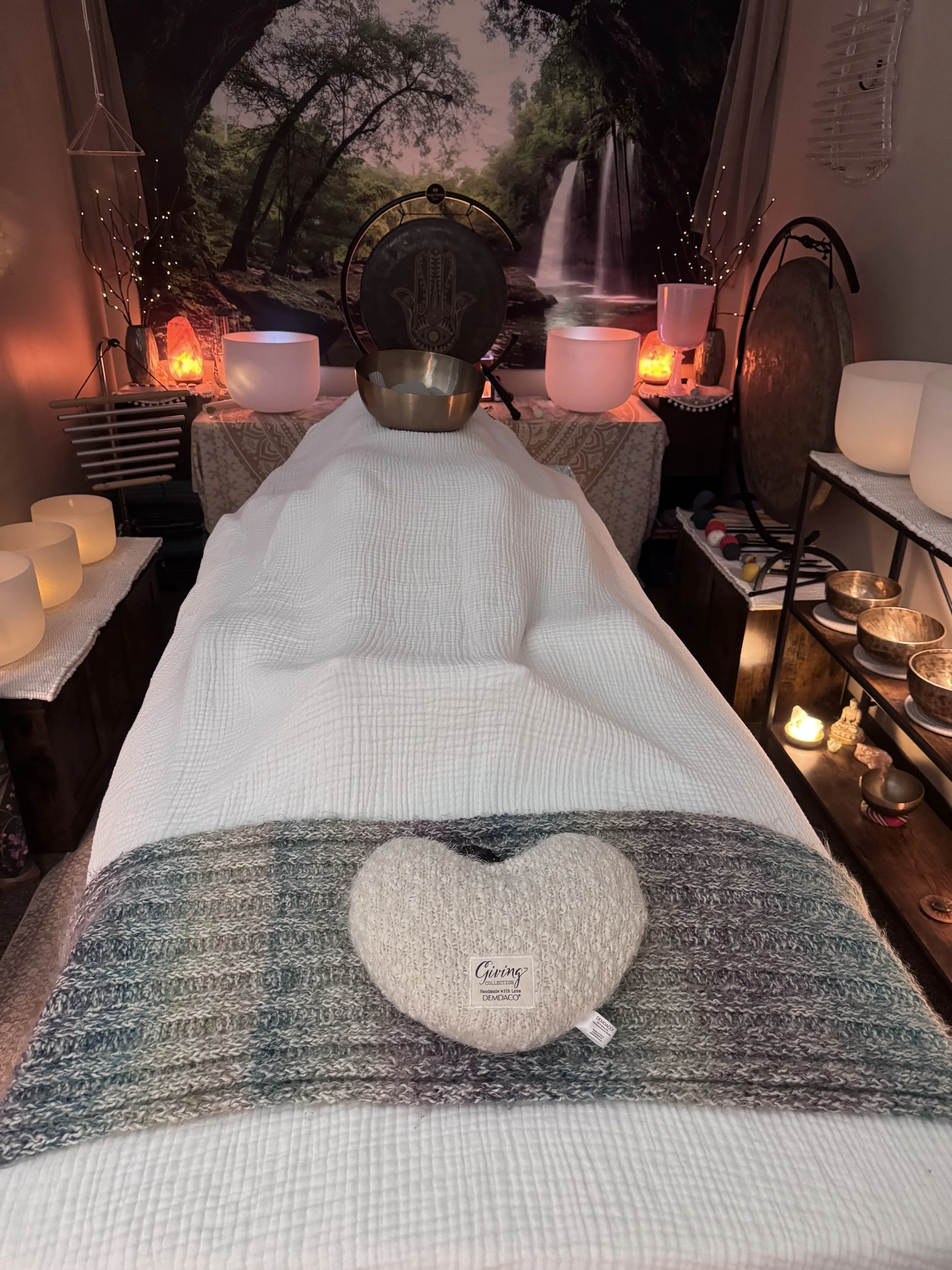 Person lying on a massage table receiving a healing treatment in a cozy room decorated with nature-themed artwork, candles, a gong, and soft lighting.