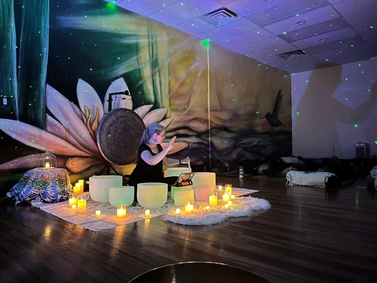 A woman practicing sound healing with crystal singing bowls in a dimly lit room decorated with candles, a large mural of a lotus flower, and a starry ceiling projection.
