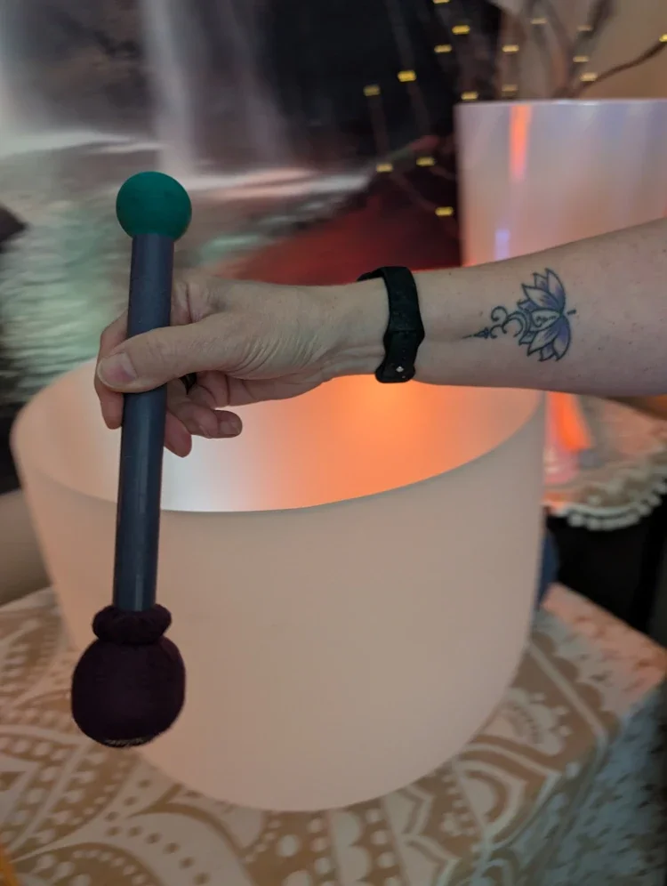 A person holding a mallet over a large singing bowl, with a tattoo of a lotus flower on their forearm.