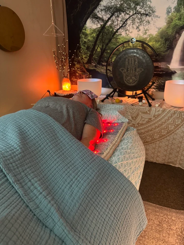 Person lying on a massage table with a blindfold, receiving a healing treatment in a cozy room decorated with nature-themed artwork, candles, a gong, and soft lighting.