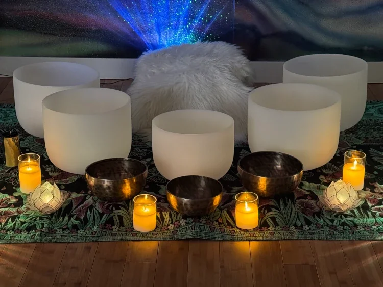 Six large white crystal singing bowls, four smaller black sound bowls, and six small lit candles on an ornate, patterned cloth.