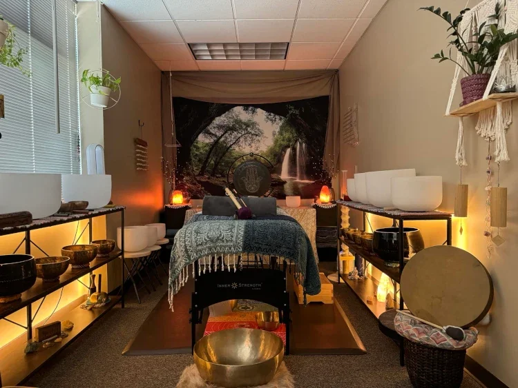 A wellness massage room with dim lighting, baskets, bowls, and candles, featuring a large waterfall and nature scene backdrop.