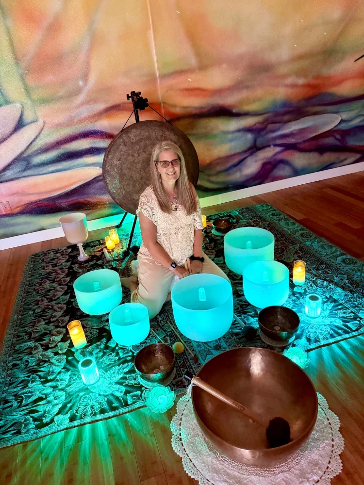 Woman sitting on a decorated mat surrounded by glowing blue and white singing bowls and candles, with a large gong and an abstract colorful mural in the background.