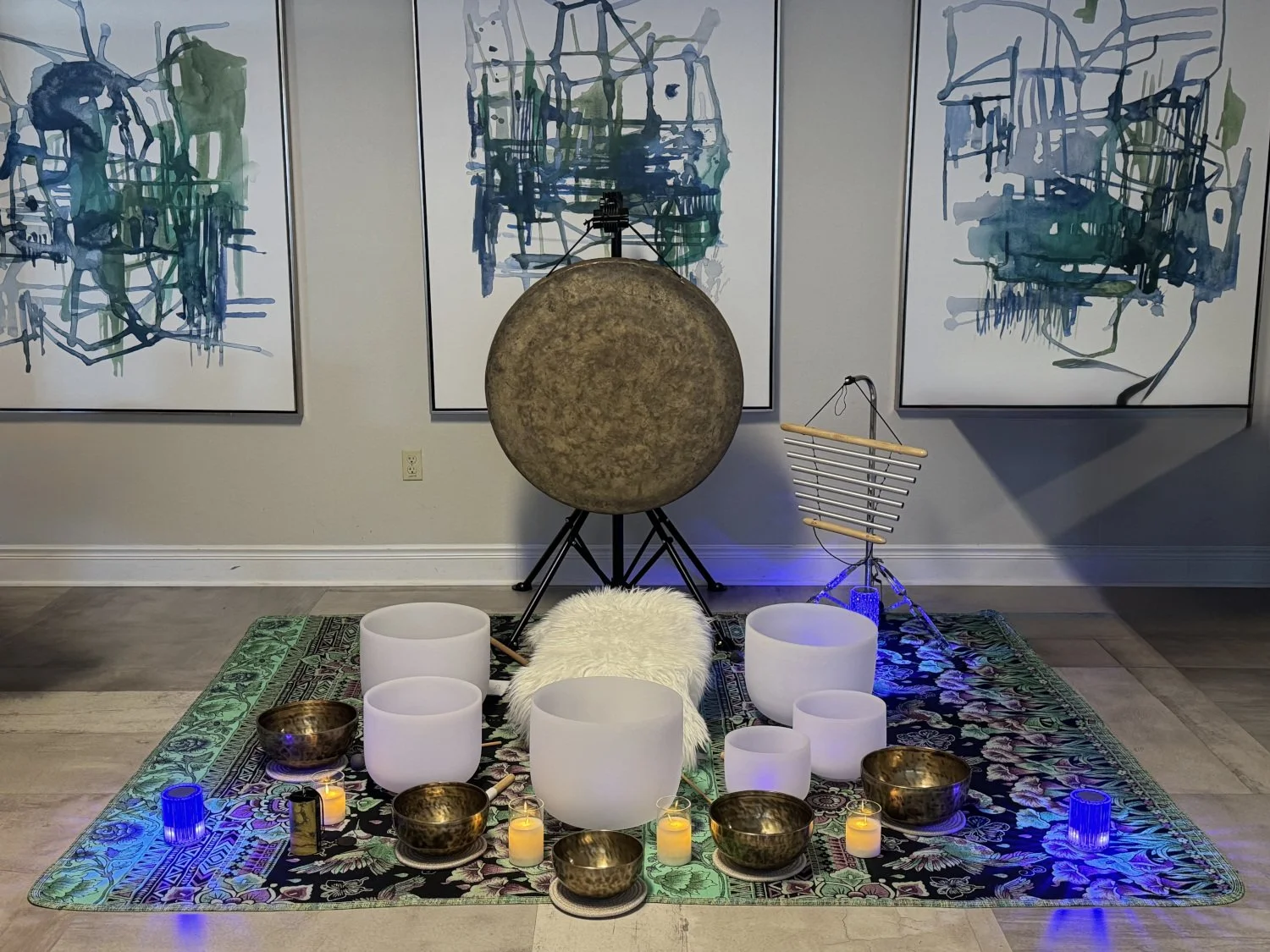 A meditation or yoga setup on a decorative rug with white crystal bowls, candles, and tibetan singing bowls surrounded by musical instruments and abstract paintings on the wall.