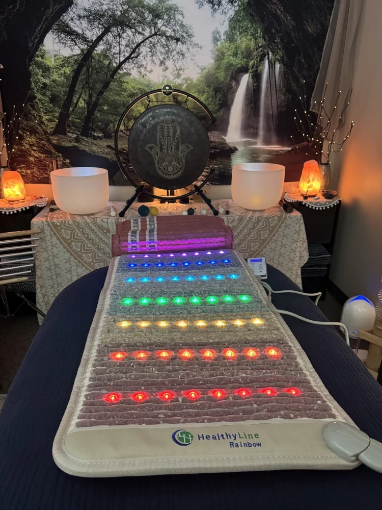 A colorful LED therapy mat with red, orange, yellow, green, blue, and purple lights arranged in parallel rows, placed on a massage table in a wellness room with nature-themed decor including a waterfall backdrop, salt lamps, and Himalayan salt night 