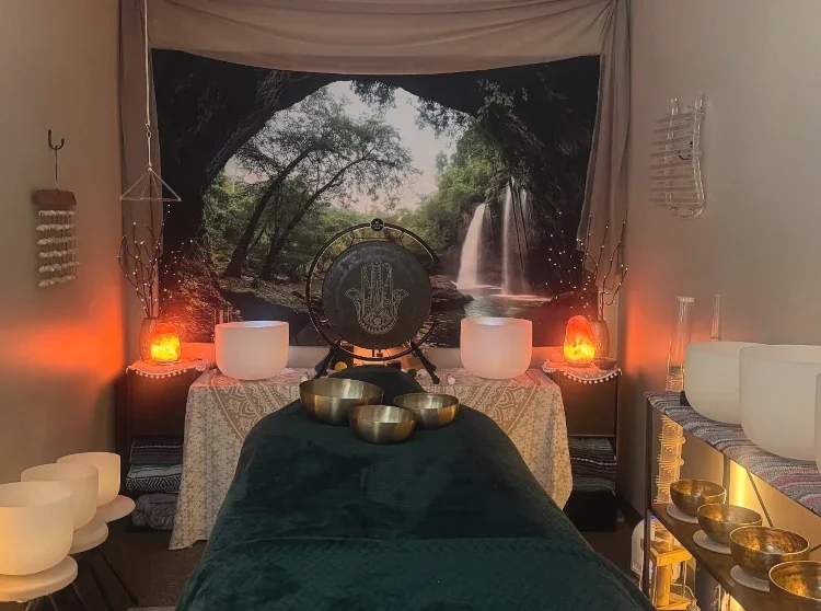 A wellness or massage room with a nature-themed backdrop of a waterfall and trees. The room features a massage table, singing bowls, salt lamps, and Himalayan salt lamps creating a calming ambiance.