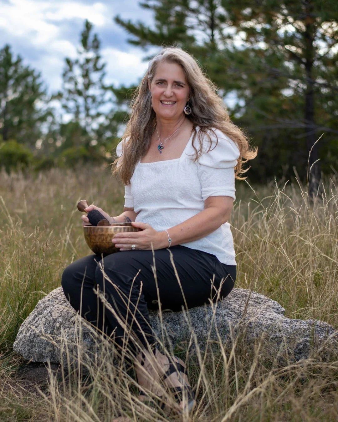 A woman with long curly hair sitting on a large rock in a grassy field, holding a singing bowl and a mallet, smiling at the camera with trees and a blue sky in the background.