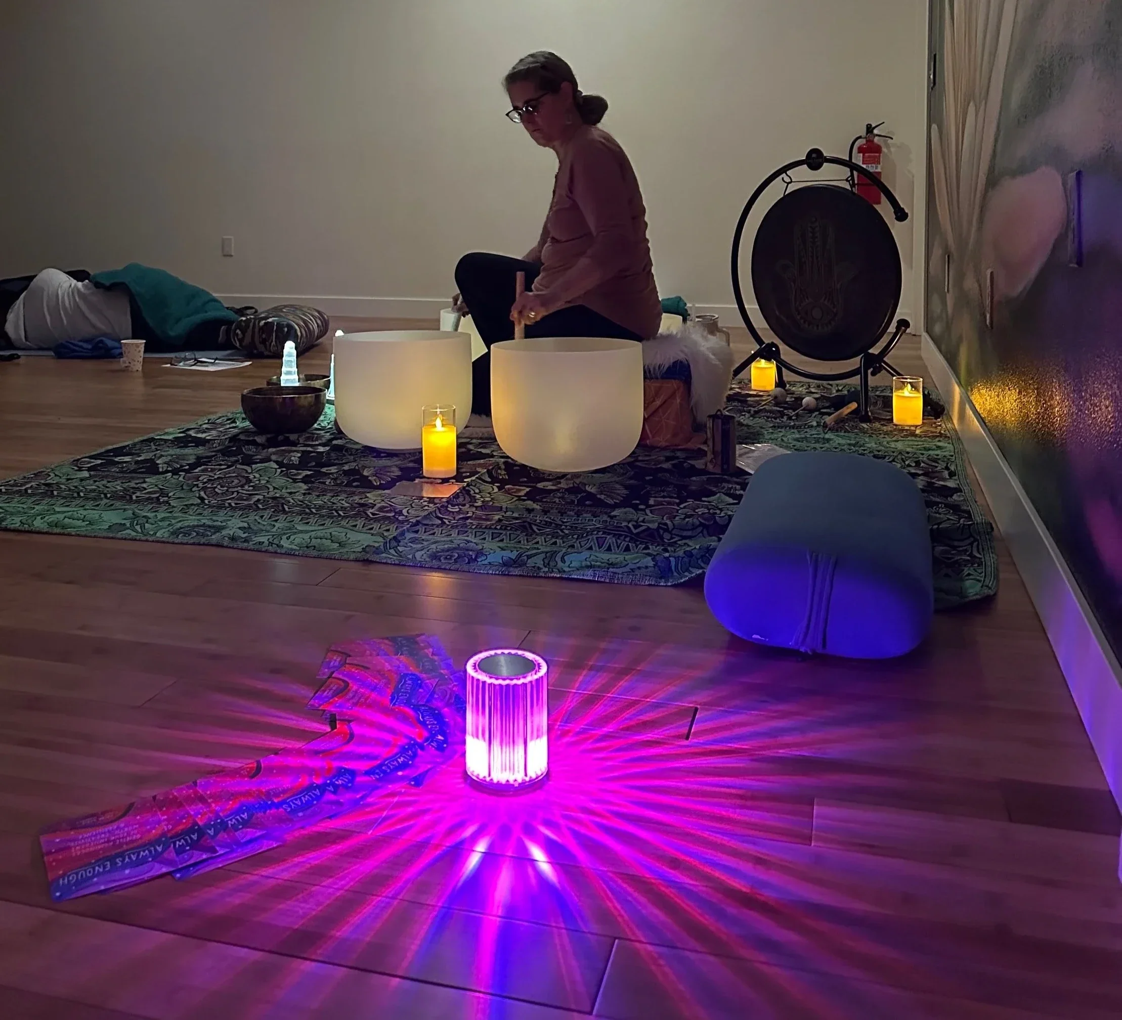 A woman practicing sound therapy with singing bowls in a room illuminated by candles and colored lights. She is seated on the floor, surrounded by a tapestry and decorative objects.