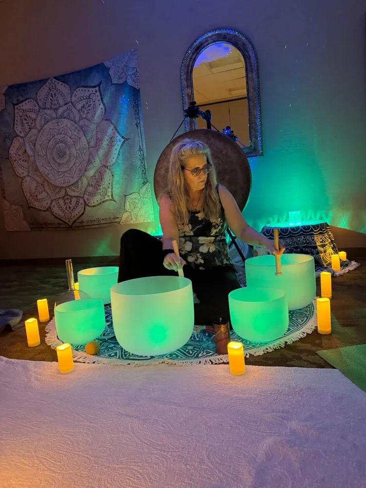 Woman sitting on the floor playing singing bowls, with glowing green and blue lights, candles, a tapestry, a mirror, and a large gong in the background.