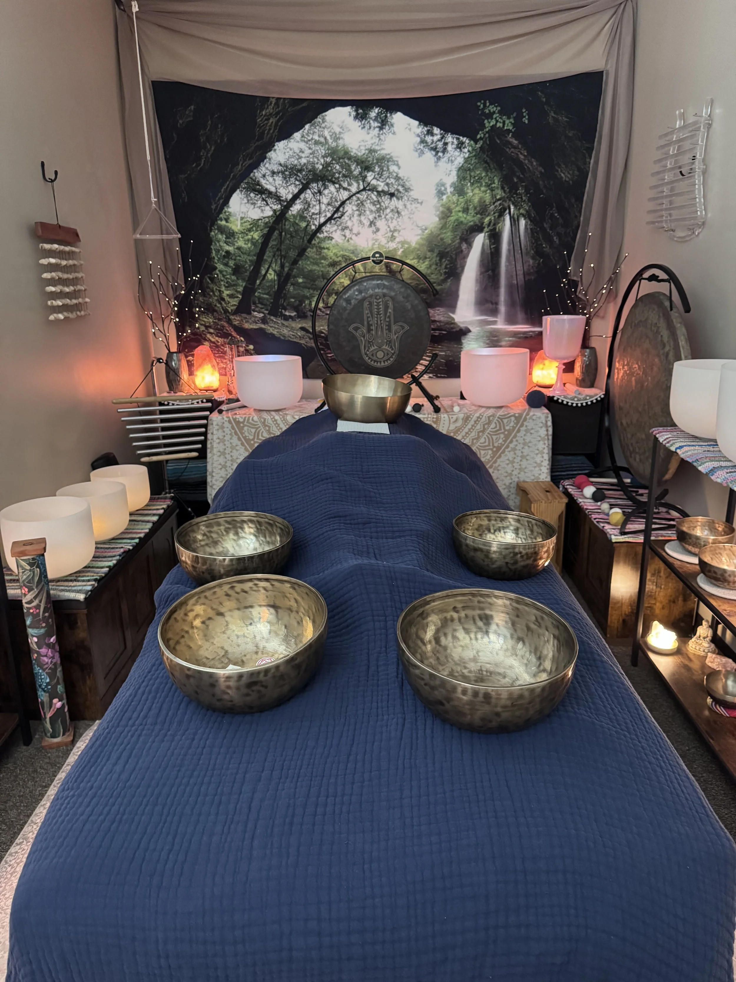 Person lying on a massage table receiving a healing treatment with Himalayan bowls on and around the body on the table in a cozy room decorated with nature-themed artwork, candles, a gong, and soft lighting.