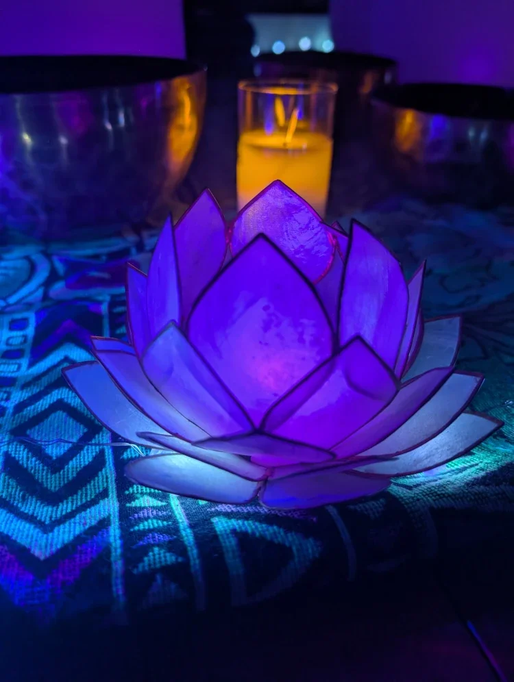 A glowing purple lotus-shaped candle holder with a lit candle inside, placed on a patterned fabric tablecloth with additional candles and metallic bowls in the background, illuminated by ambient purple and yellow lighting.