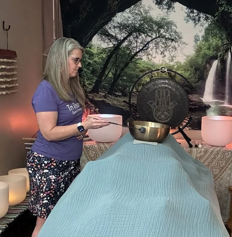 A woman with long blonde hair, wearing glasses, a purple t-shirt, and patterned shorts, standing in a room with a nature-themed mural, playing a singing bowl with a mallet.