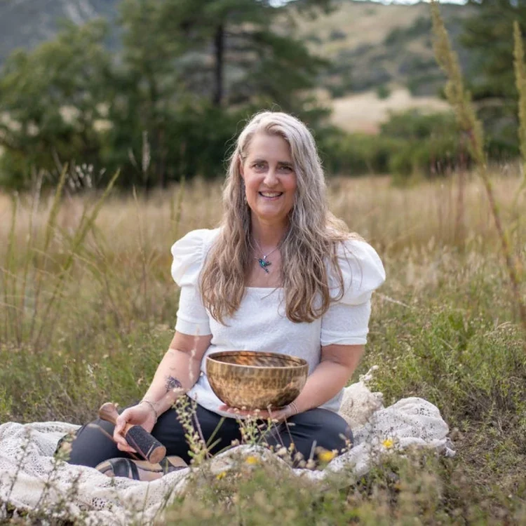 A woman with long blonde hair, wearing a white top, sitting on a blanket in a grassy field, holding a wooden singing bowl and a mallet, smiling at the camera.
