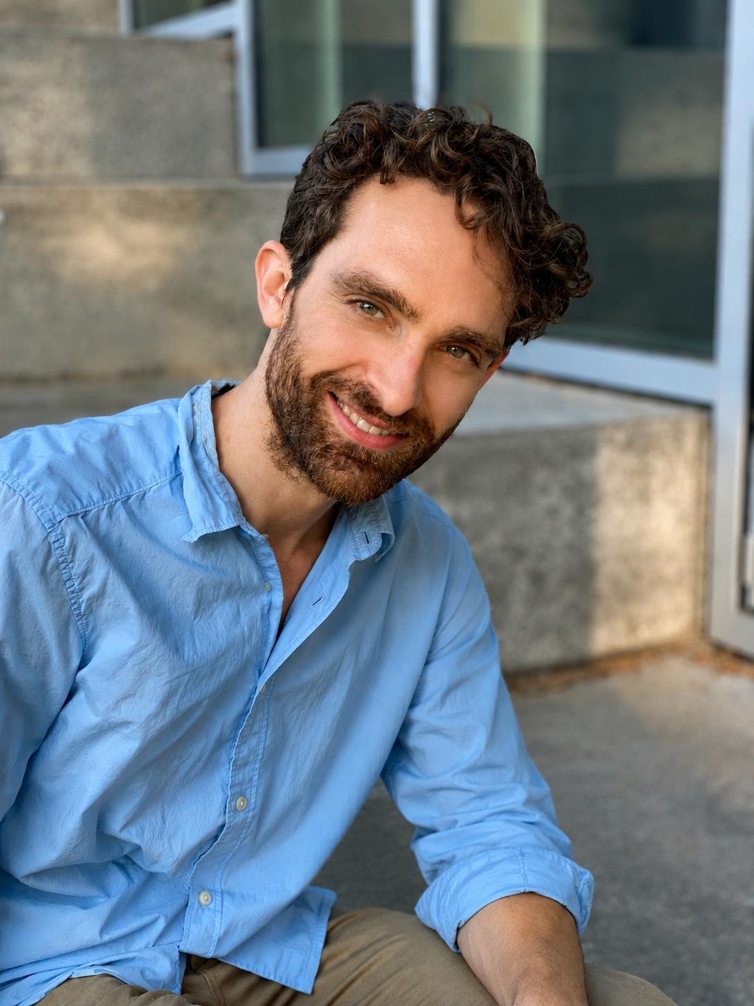A smiling man with curly brown hair and a beard wearing a light blue button-up shirt, sitting outside near some steps and glass doors.