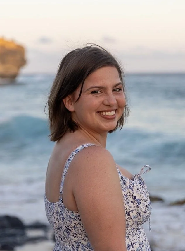 A young woman with shoulder-length brown hair smiling at the camera, standing on a beach with the ocean and a rocky cliff in the background.