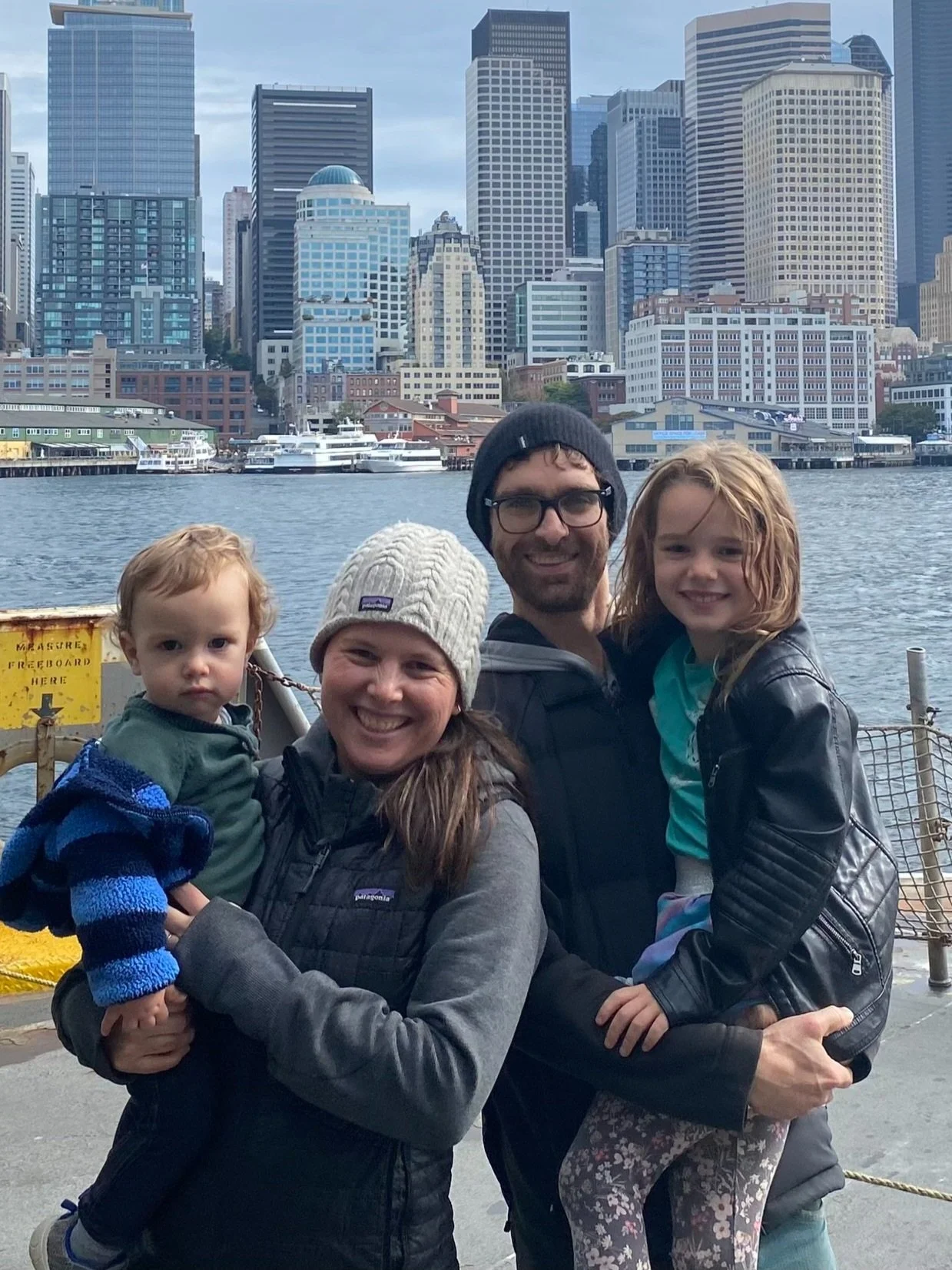 A family of four is posing on a waterfront with a city skyline in the background. The family includes a woman holding a young boy, a man, and a young girl sitting on the man’s lap. They are dressed warmly and smiling, with boats on the water and tall buildings behind them.