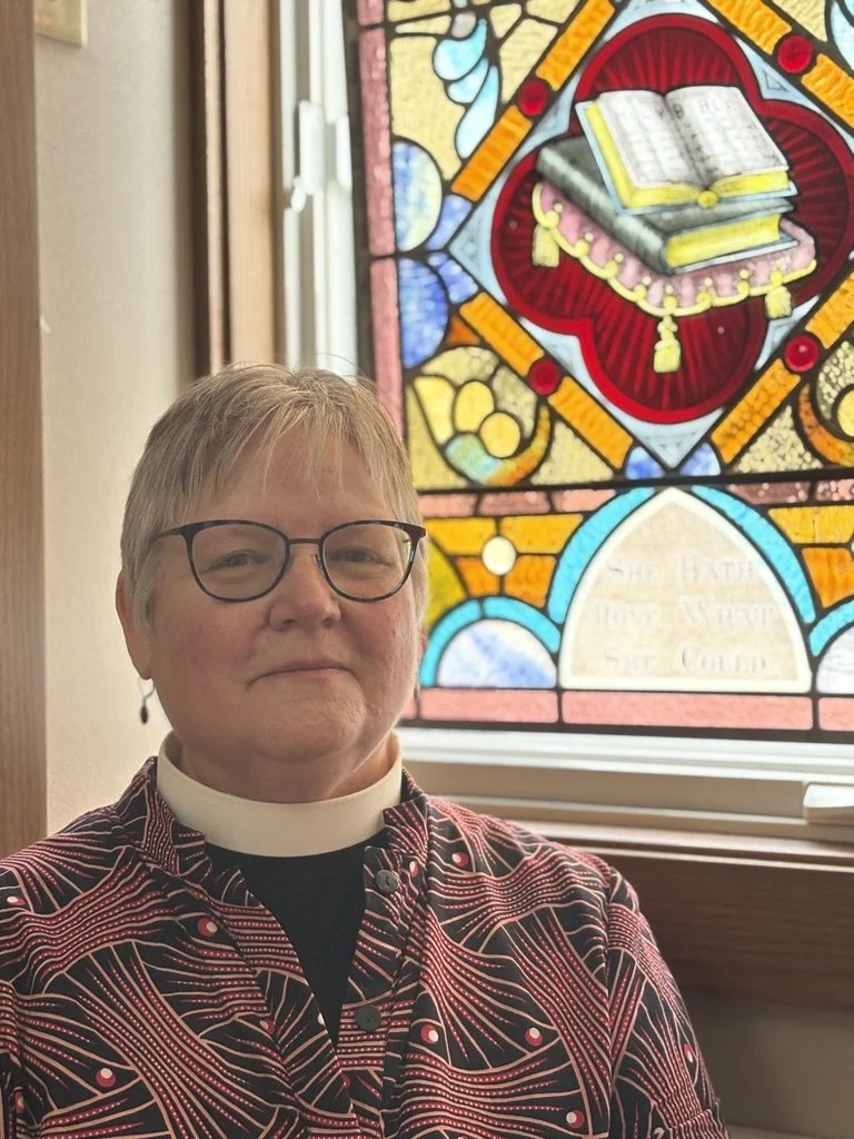 Helen Svoboda-Barber in clerical collar seated in front of a colorful stained glass window