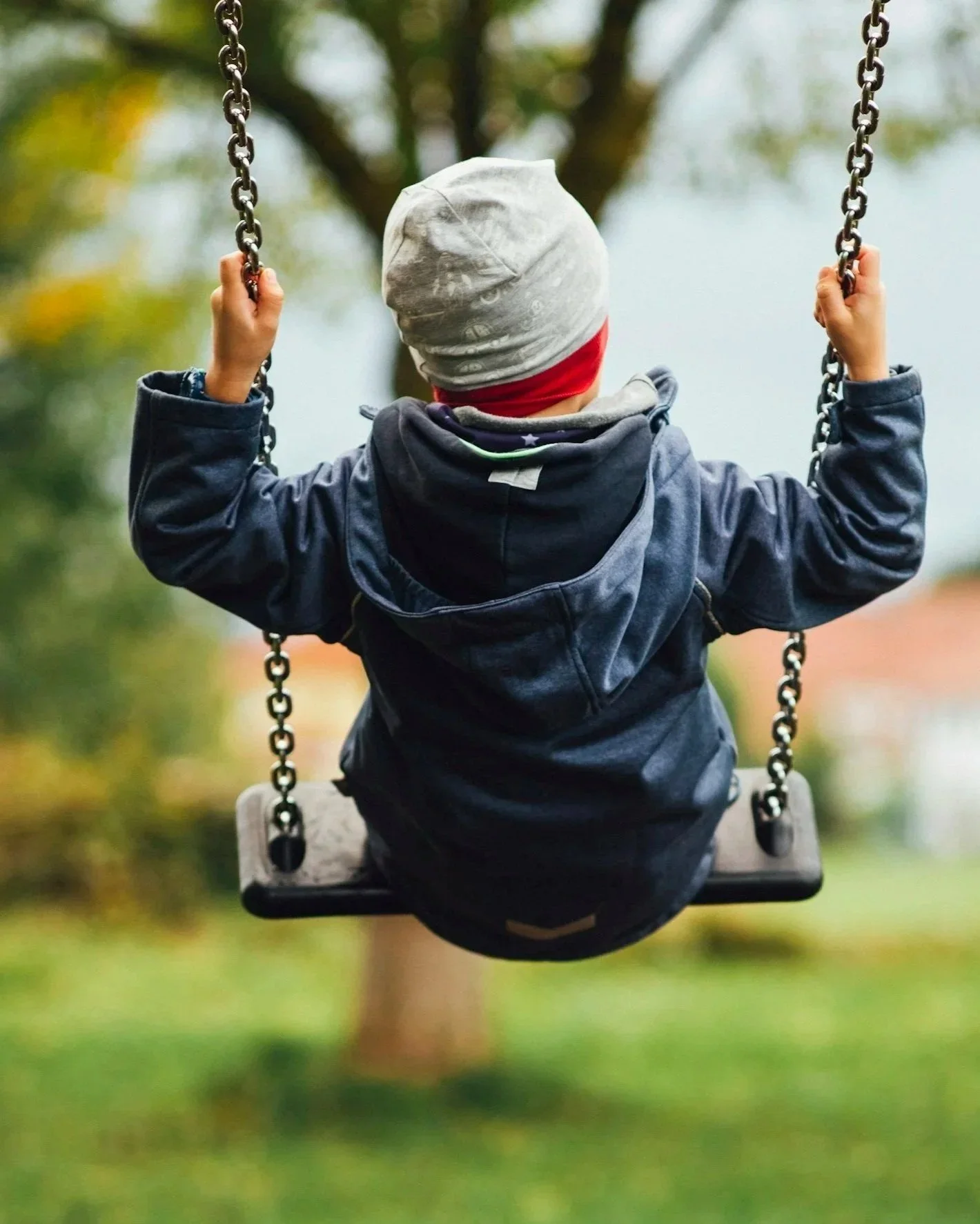 A young boy wearing a gray beanie and dark jacket is sitting on a swing outdoors, holding the chains with both hands, with trees and blurred foliage in the background.