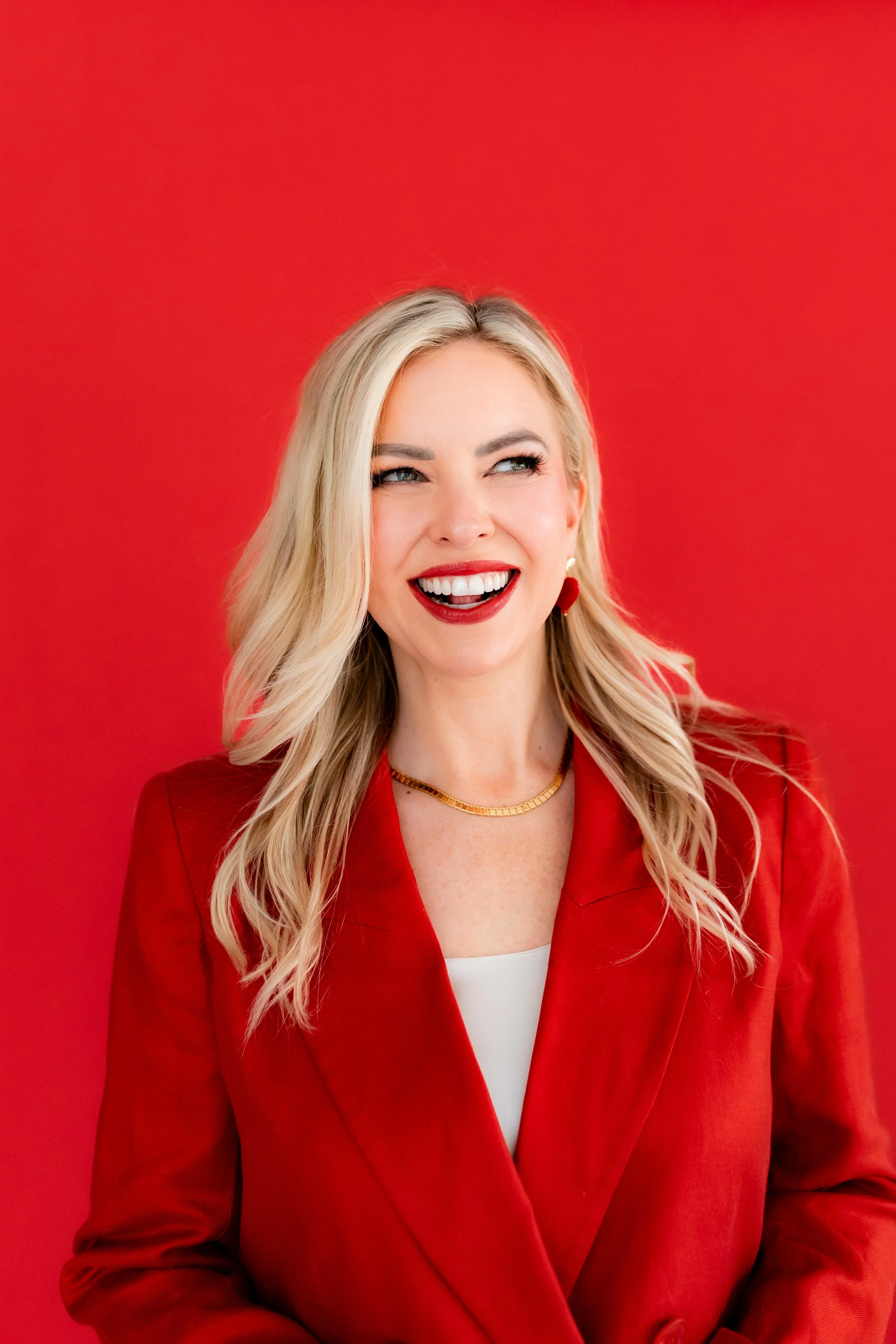 Monica Gostkiewicz smiling and looking to the side, wearing a red blazer, white top, gold necklace, earrings, and red lipstick, standing against a red background.