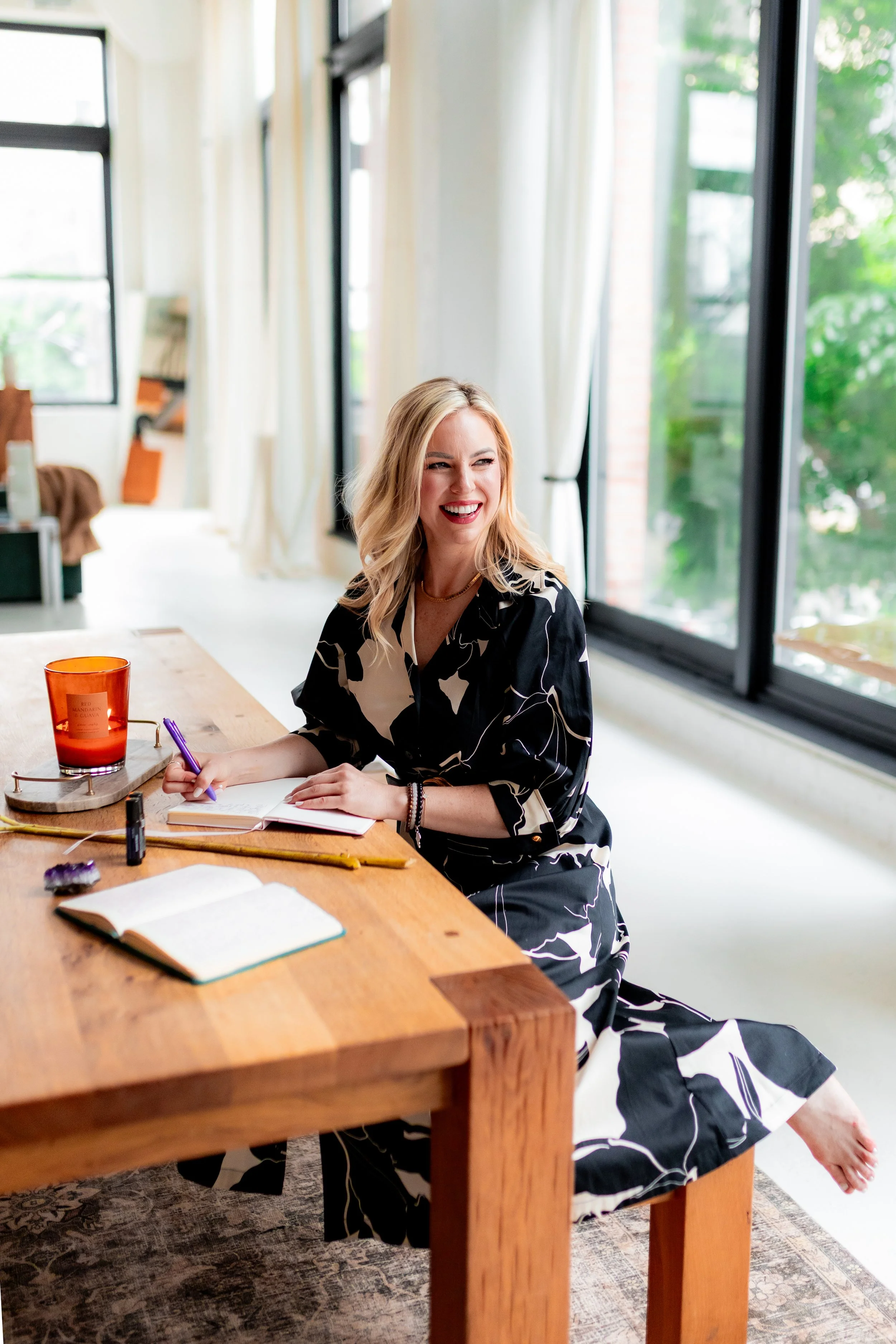 A woman with blonde hair in a black and white dress sitting at a wooden table, writing in a notebook, smiling, near large windows with white curtains, in a bright room.