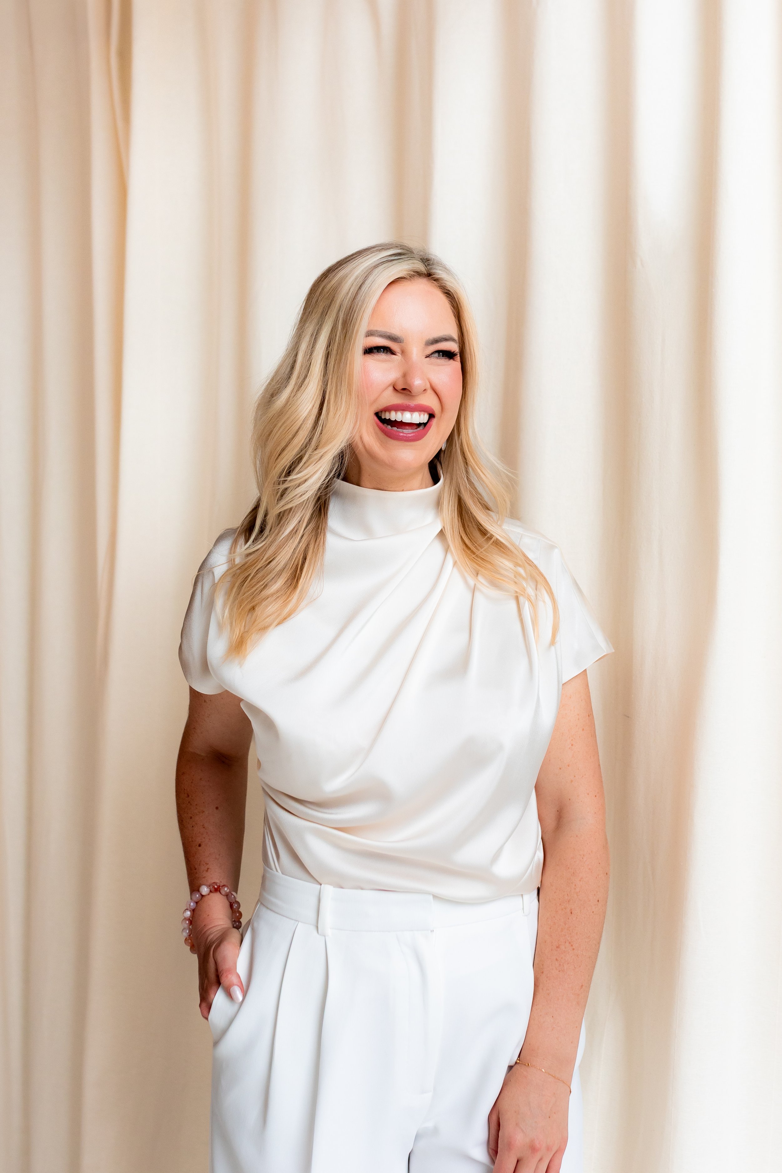 Monica Gostkiewicz smiling, dressed in a white top and pants, standing in front of beige curtains.