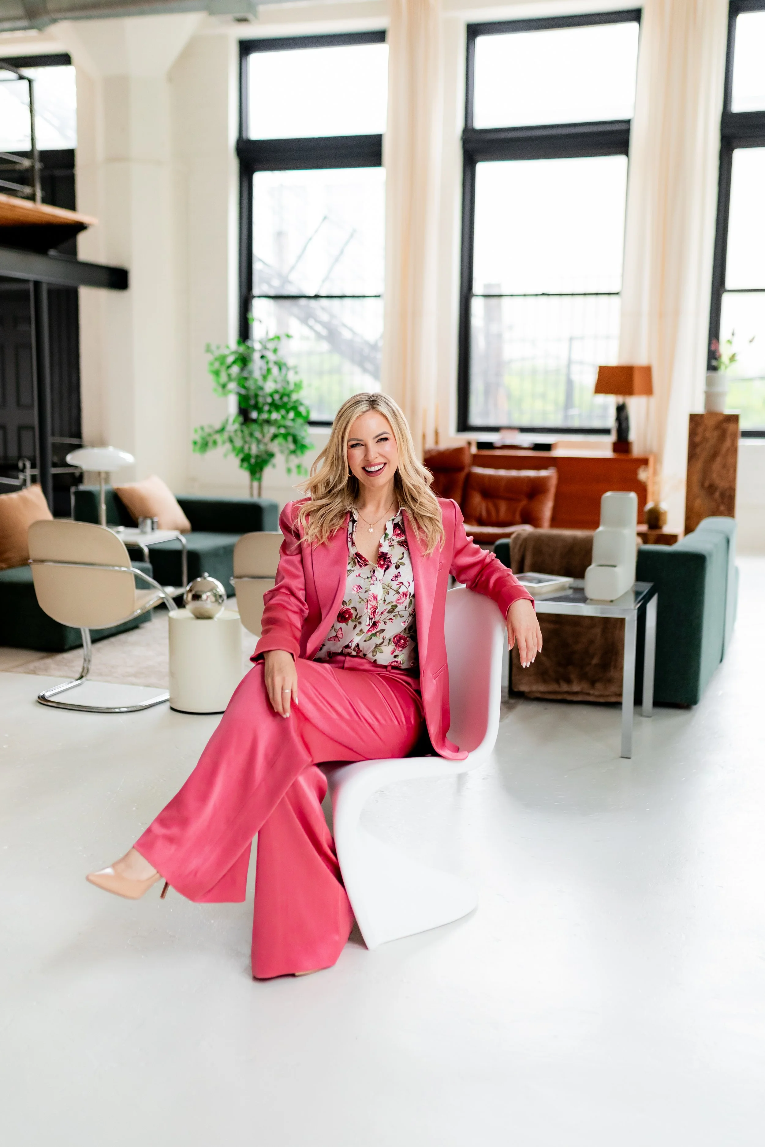 Monica Gostkiewicz in a pink suit sitting on a white modern chair in a brightly lit room with large windows, plants, and contemporary decor.