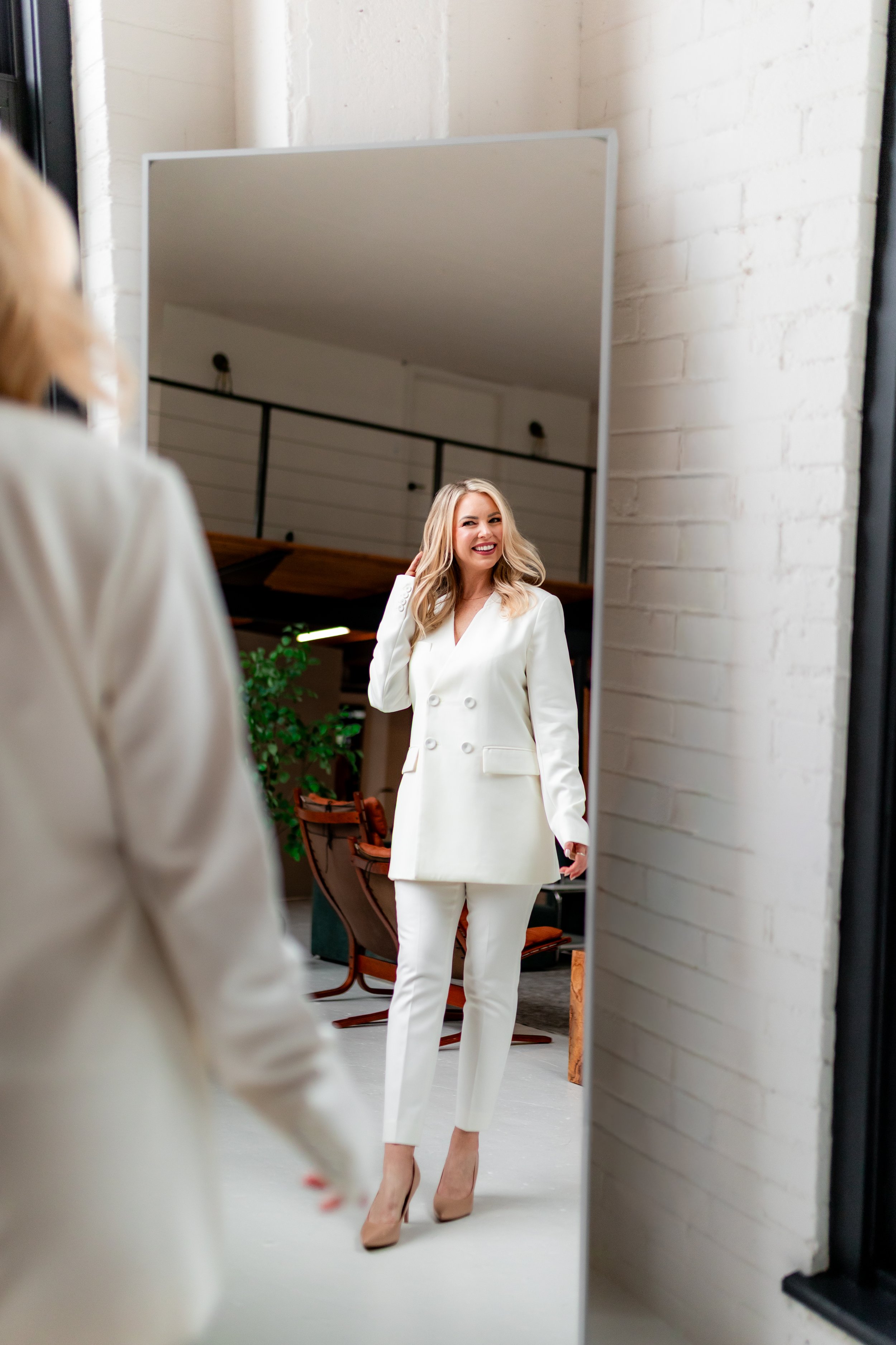 Monica Gostkiewicz in a white suit looking at her reflection in a full-length mirror, with a white brick wall and a plant in the background.