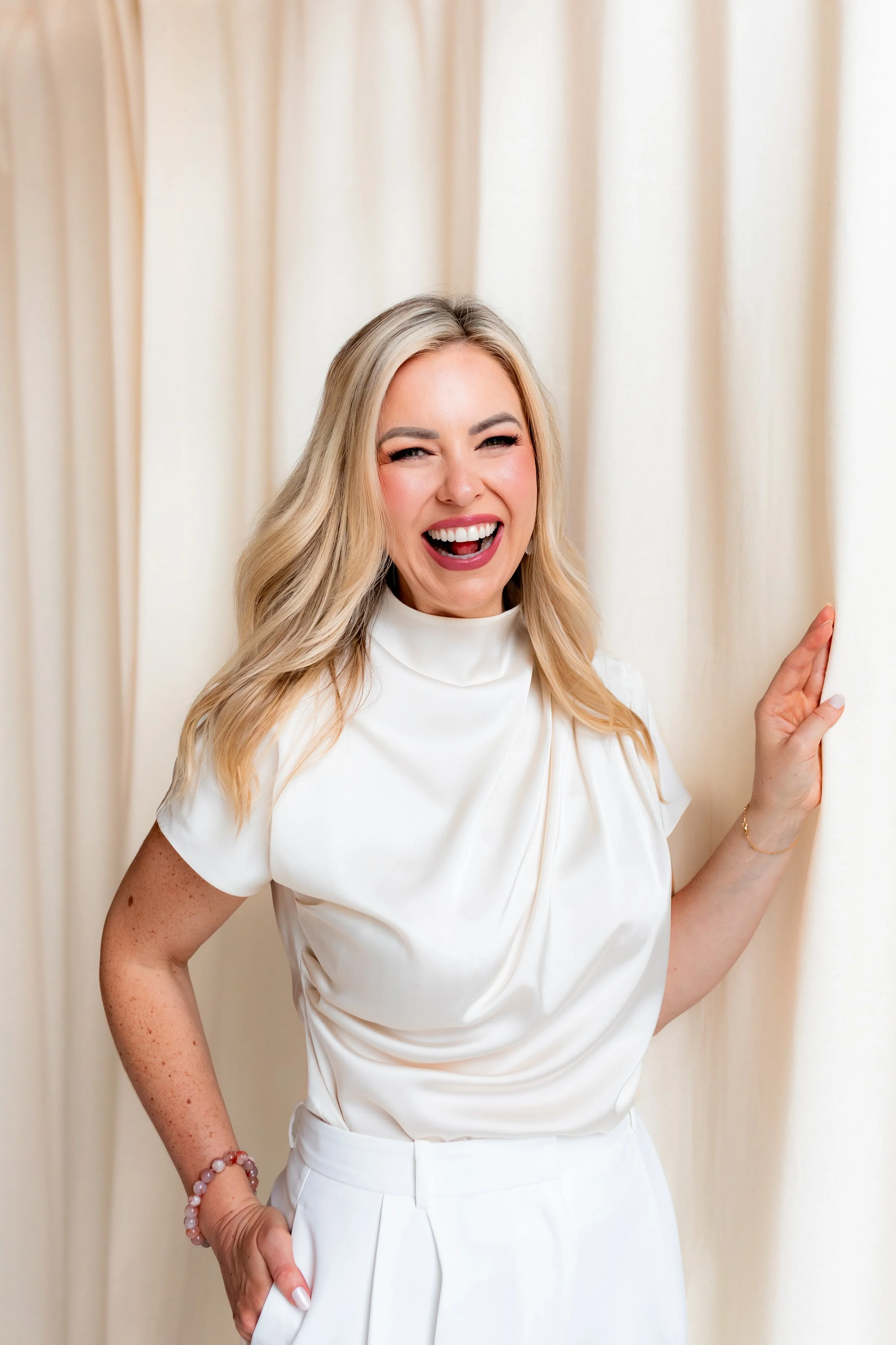 A woman with long blonde hair smiling and looking at the camera, wearing a white short-sleeved top and white pants, standing in front of light-colored curtains.
