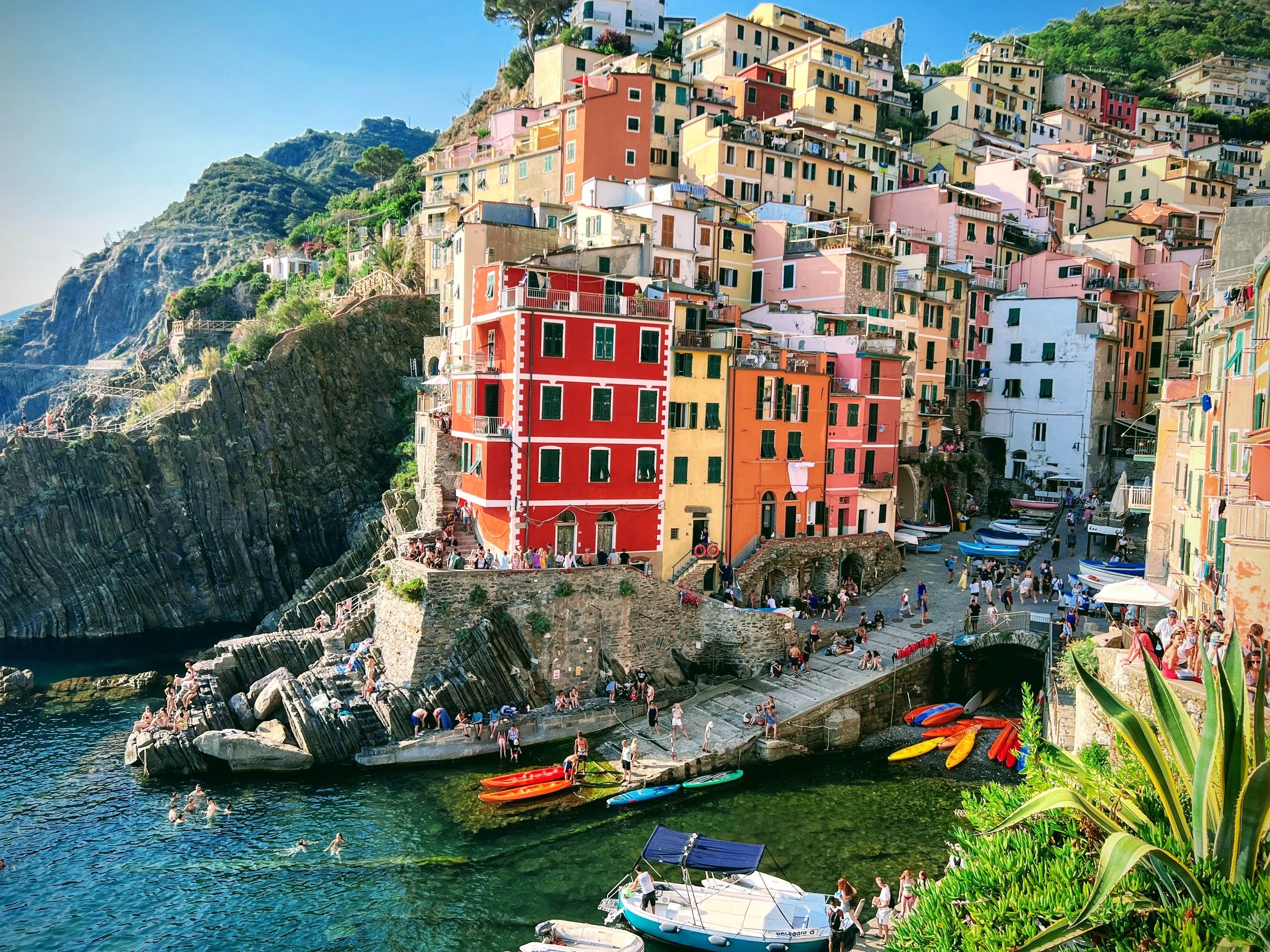 Colorful houses on a hillside overlooking the marina in Cinque Terre, Italy, with boats and kayaks in the water and tourists on the rocks and dock.