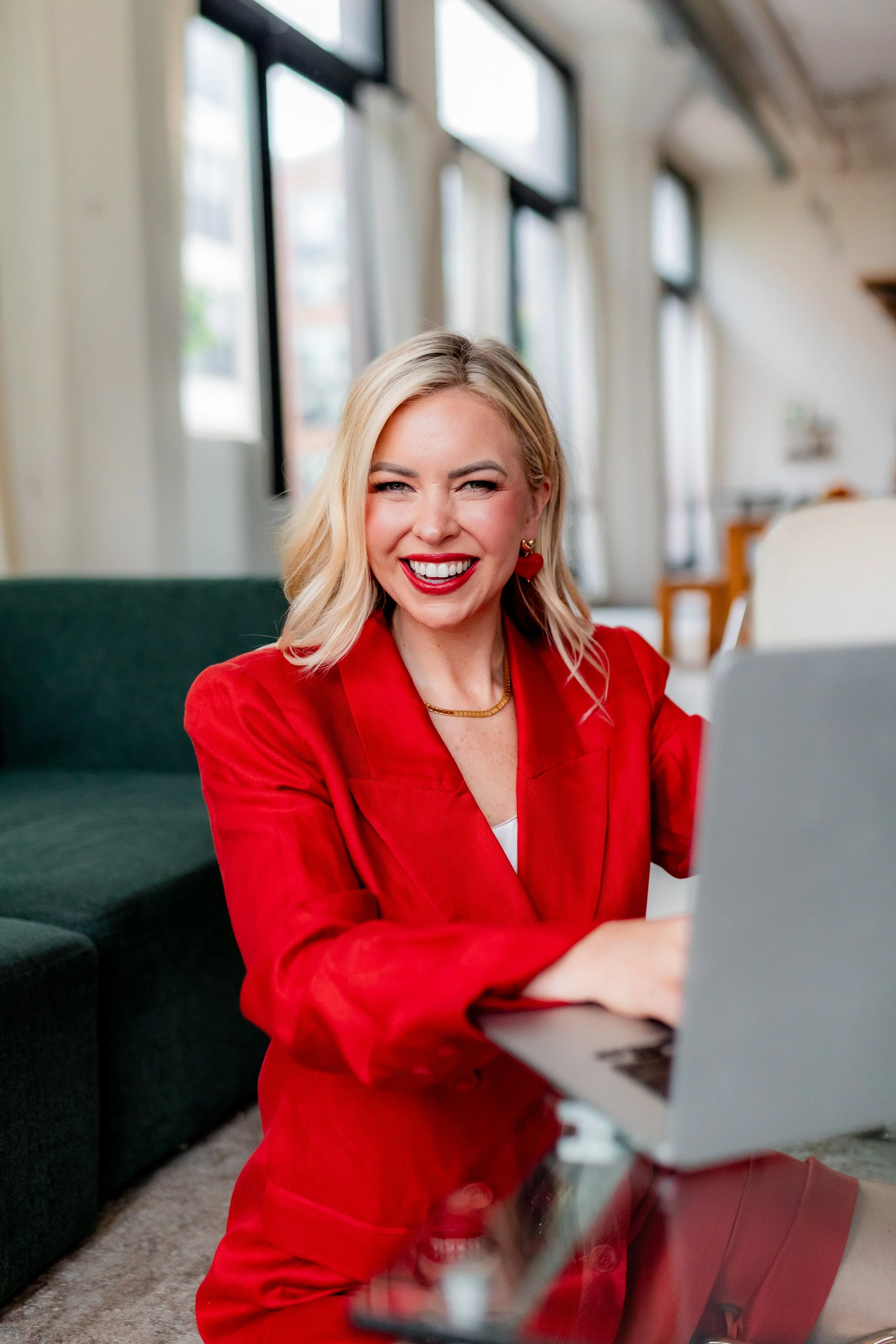 A woman with blonde hair, wearing a red blazer and dress with red lipstick, sitting on a green sofa in a bright room with large windows, smiling while looking at a laptop.