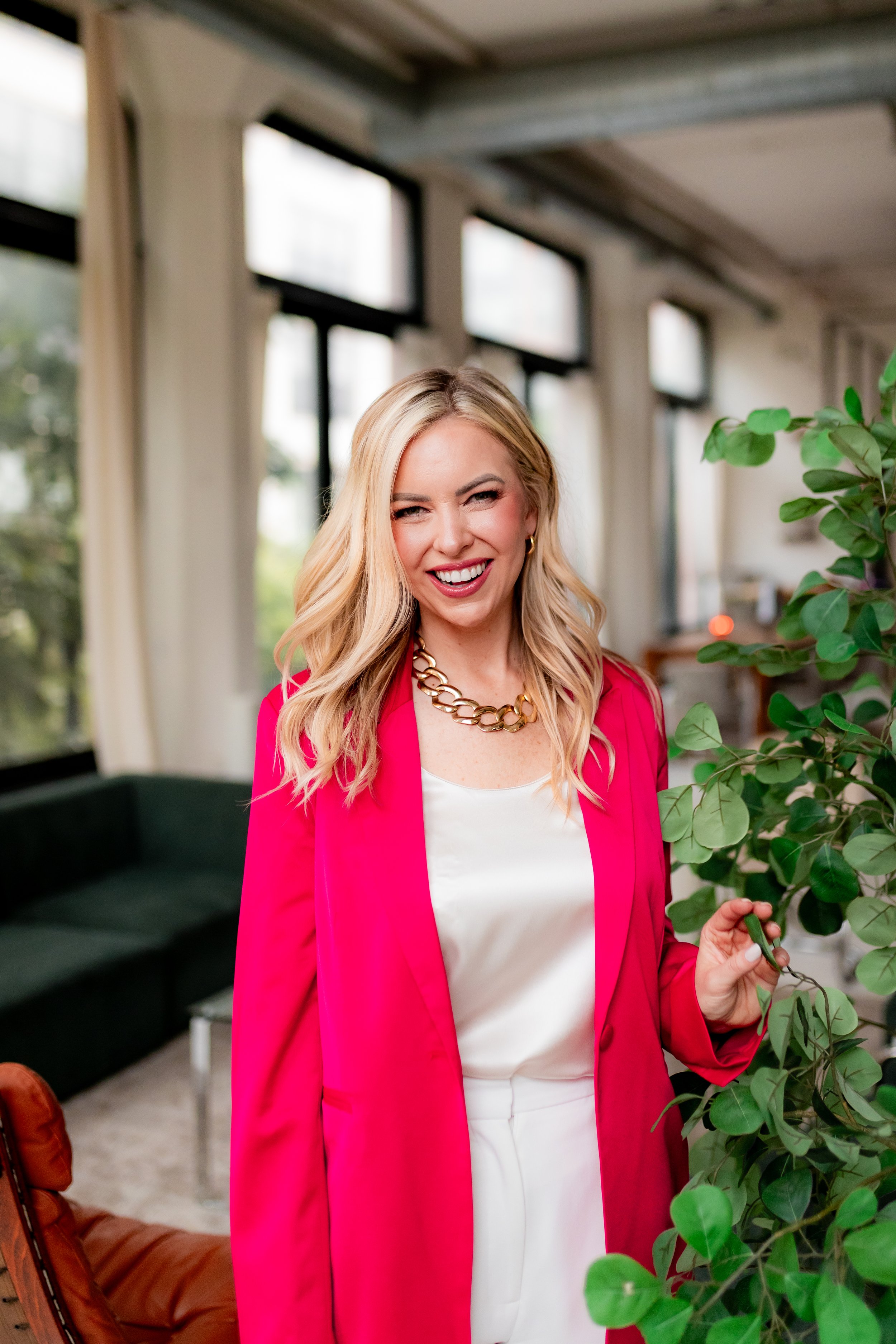 A woman with blonde hair wearing a pink blazer, white top, and gold necklace, smiling and standing next to a green potted plant in an indoor setting with large windows.