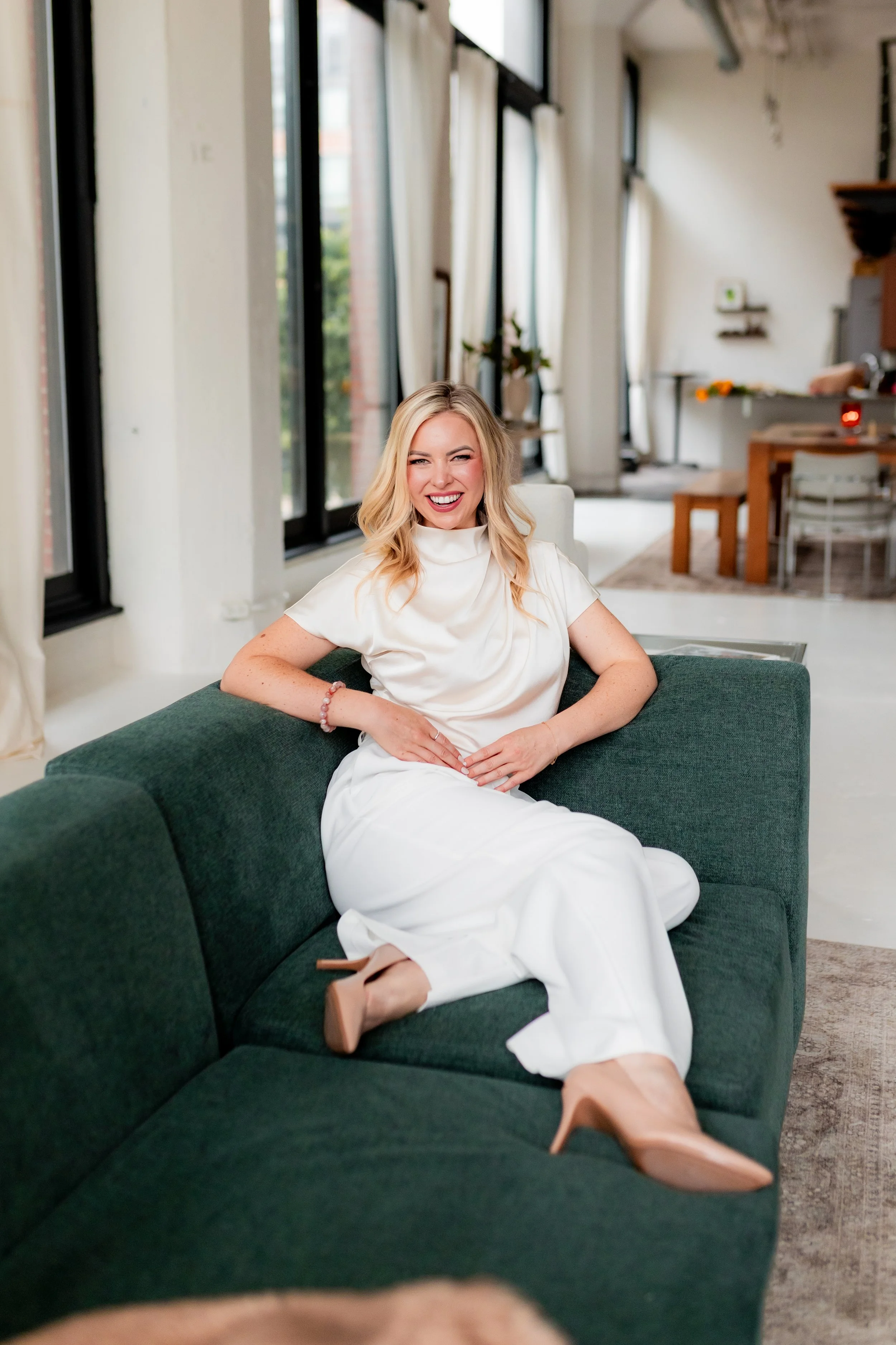 A woman with blonde hair, wearing a white satin blouse and white pants, sitting on a green sofa in a well-lit room, smiling and laughing.