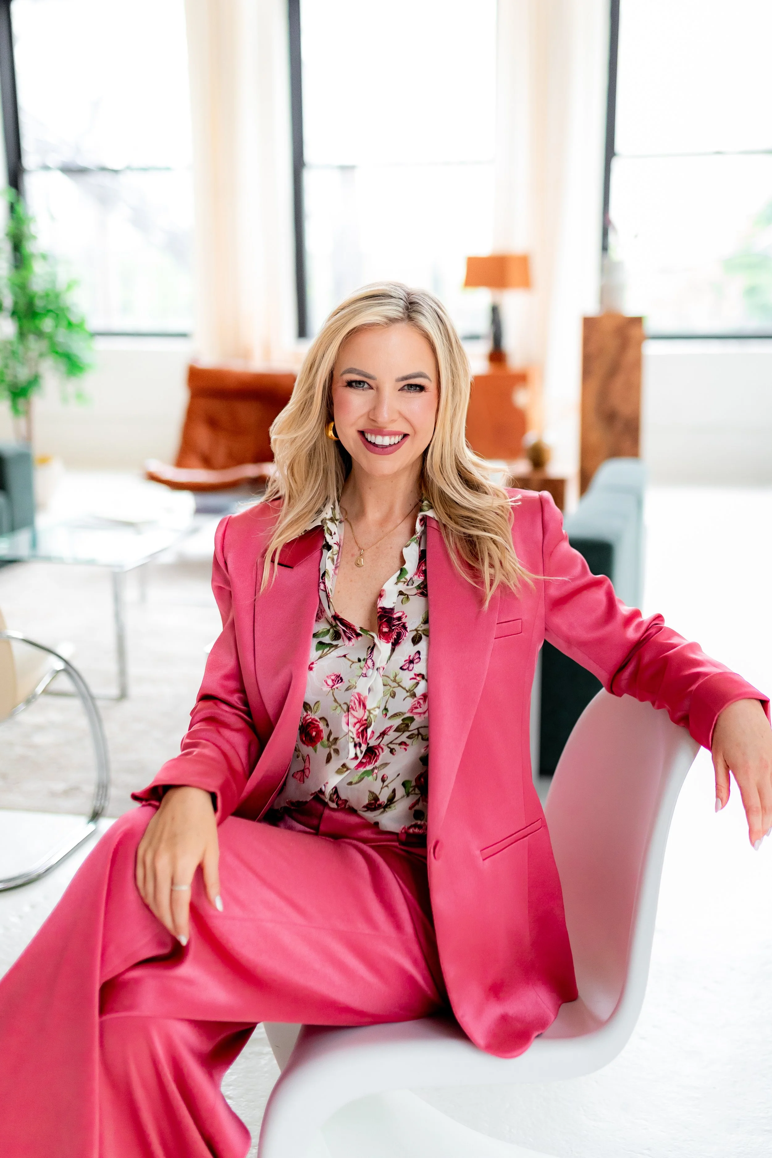 A woman with blonde hair wearing a pink suit and floral blouse sitting in a modern, brightly lit room with large windows, smiling at the camera.