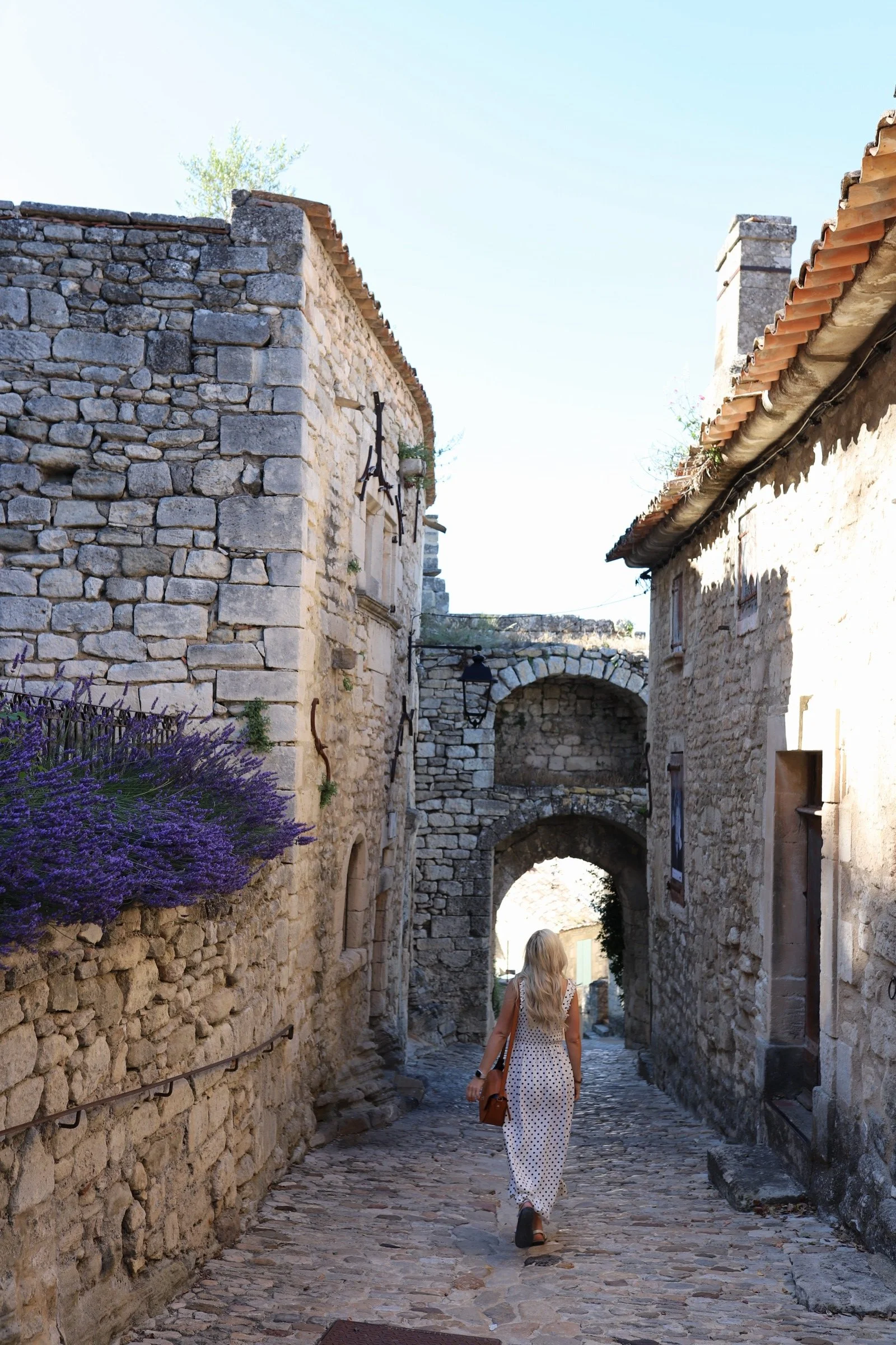 A woman with long blonde hair wearing a white polka dot dress and brown handbag walking down a cobblestone street between old stone buildings with archways, a street lamp, and purple flowers on the left.