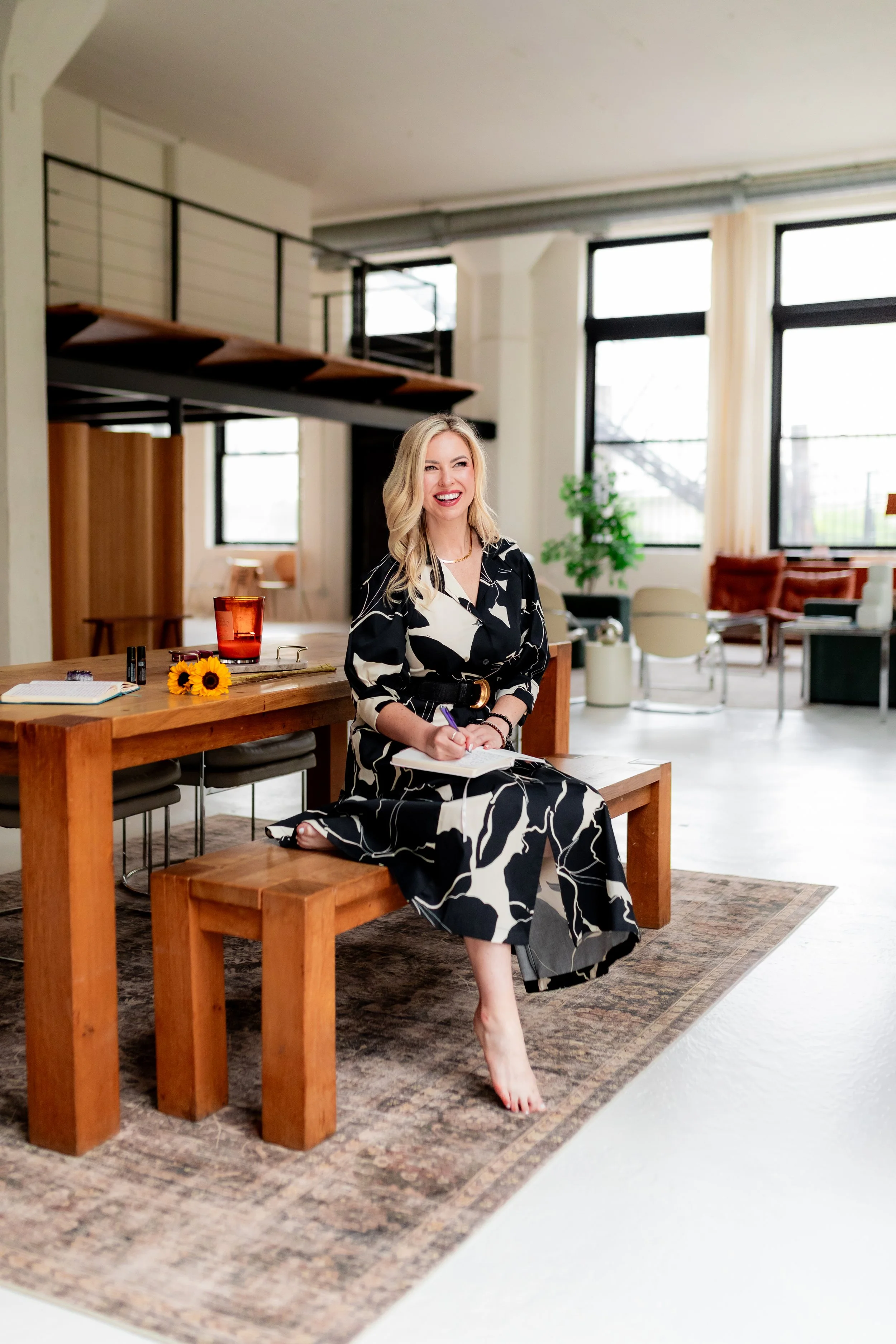Monica Gostkiewicz sitting on a wooden bench in a modern, well-lit indoor space, smiling, with her bare feet touching a patterned rug.