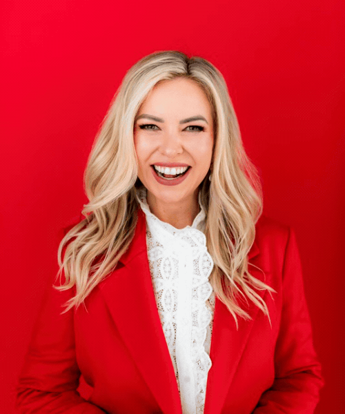 Woman with blonde hair smiling and wearing a red blazer and white lace blouse against a red background.