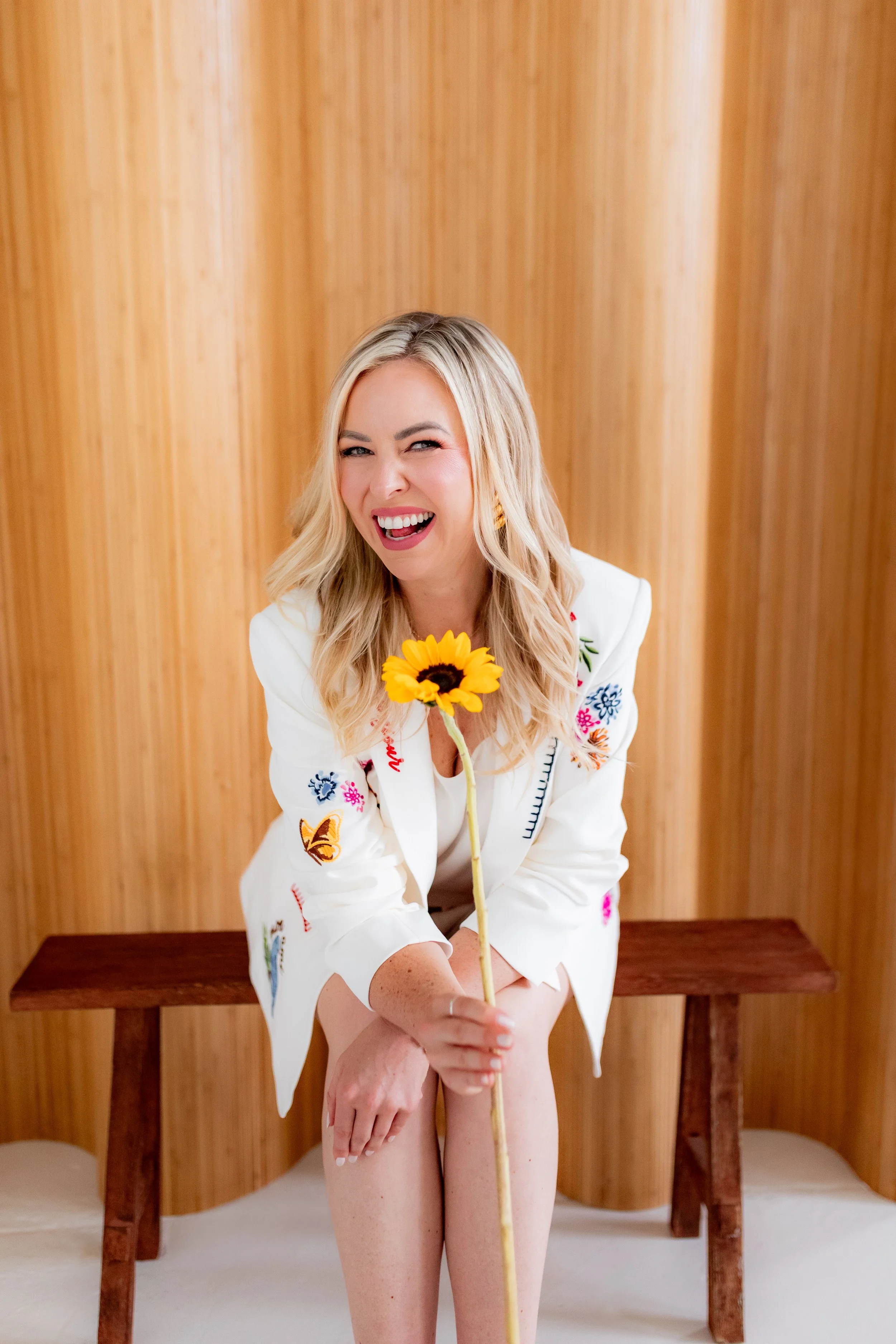 Monica Gostkiewicz wearing a white embroidered jacket, smiling and winking, holding a sunflower in her hand, sitting on a wooden bench in front of a wood-paneled wall.