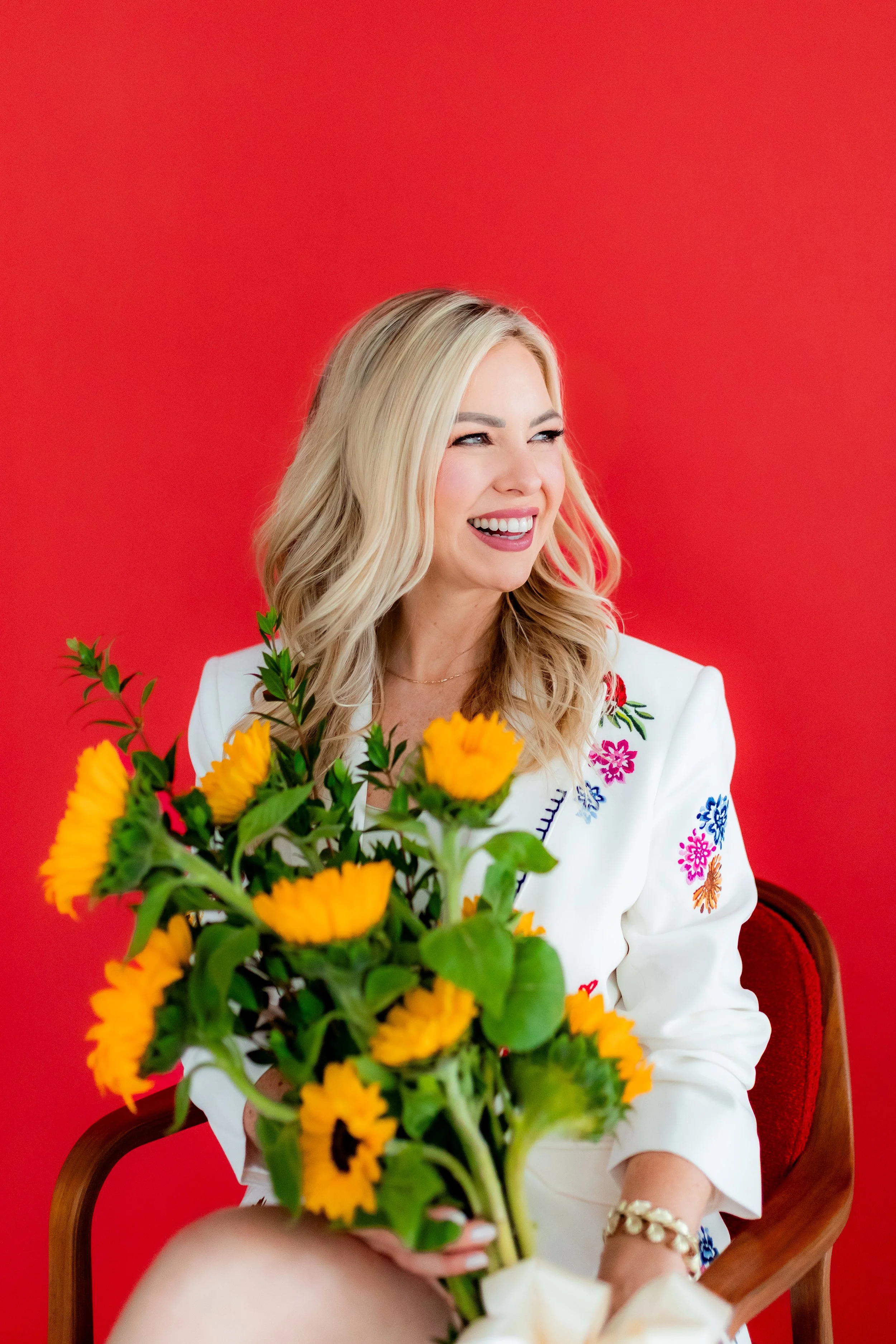 Monica Gostkiewicz smiling while holding a bouquet of yellow flowers, sitting on a red chair in front of a red background, wearing a white embroidered blouse.