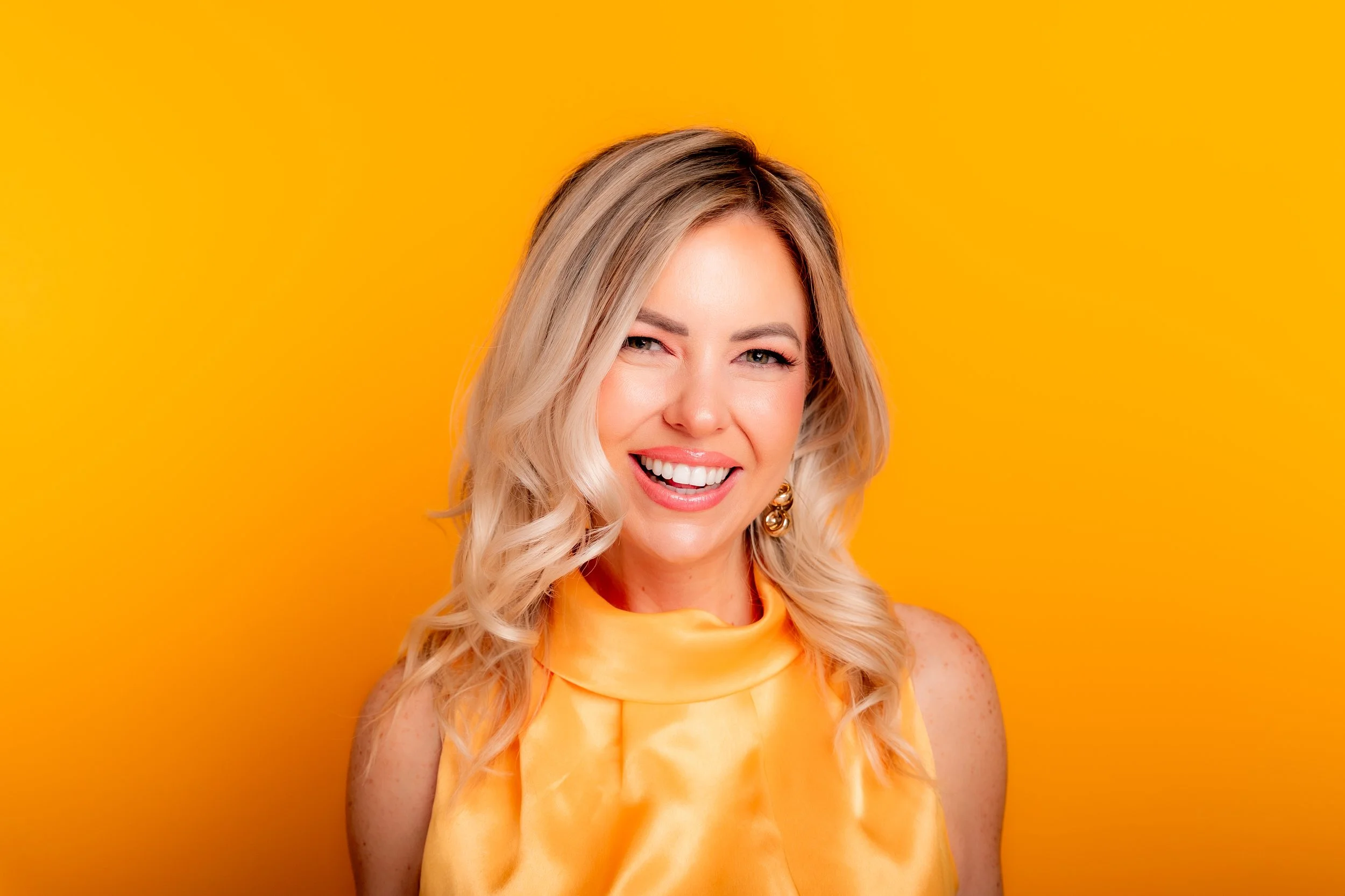 Monica Gostkiewicz smiling and winking, wearing an orange outfit, against a bright yellow background.