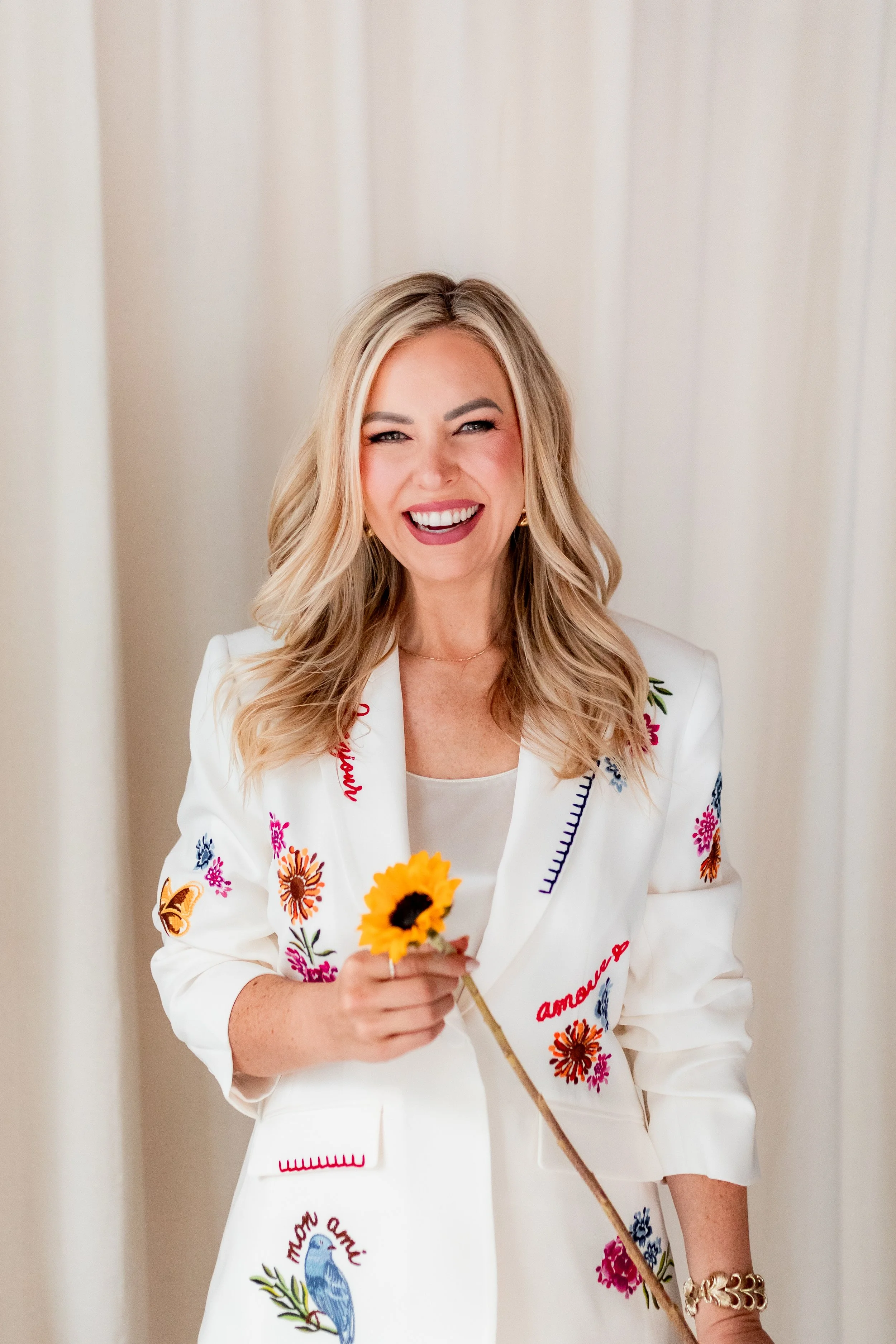 Monica Gostkiewicz wearing a white embroidered jacket with colorful floral and bird designs, holding a sunflower, standing against a plain background.