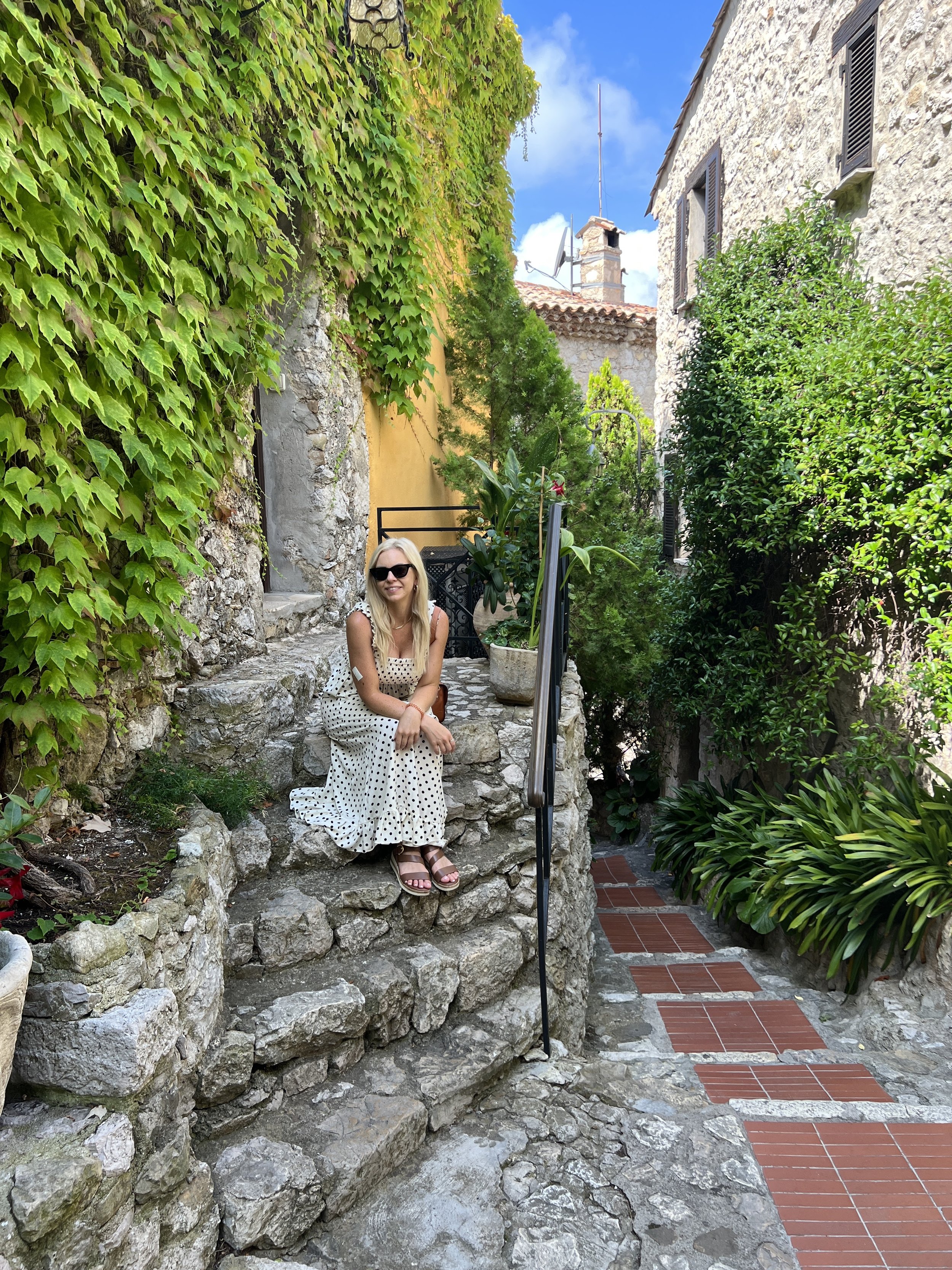 A woman with blonde hair, wearing sunglasses and a white polka dot dress, sitting on a stone ledge in an outdoor garden. The garden has green ivy on the left wall, various plants and shrubs on the right, and a stone pathway with red bricks. She is smiling and the sky is partly cloudy with blue and white clouds.